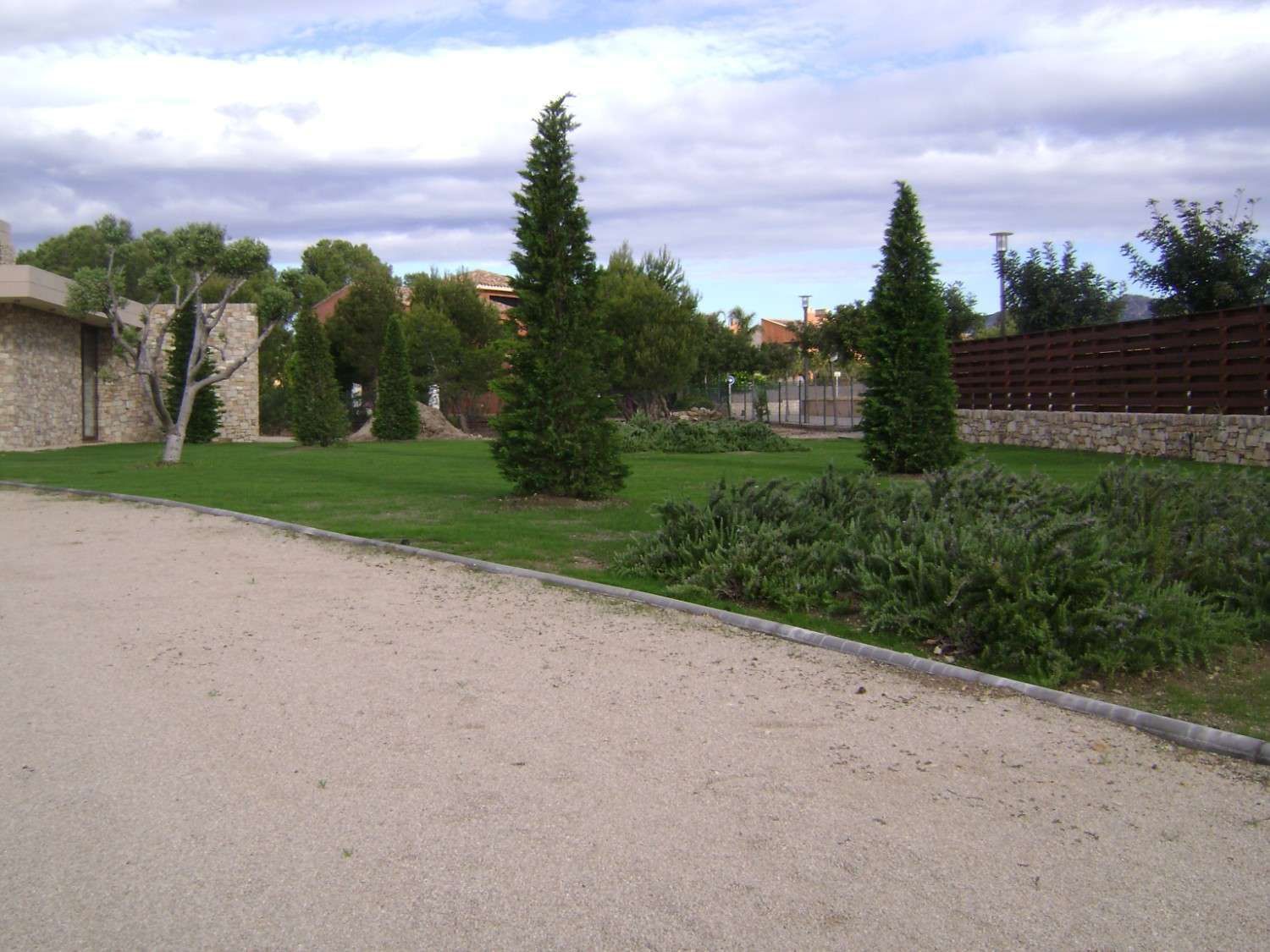Sendero de grava junto a césped verde, arbustos y altas coníferas bajo un cielo nublado.