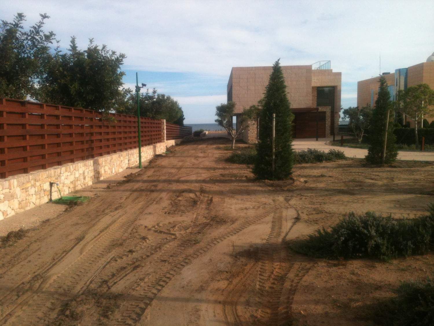 Sendero arenoso entre edificios y una valla, con árboles y arbustos bajo un cielo nublado.