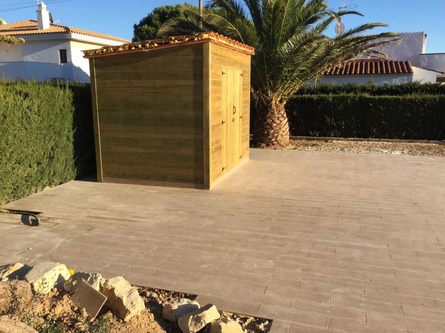 Caseta de madera en un patio trasero pavimentado con setos, una palmera y una casa al fondo.