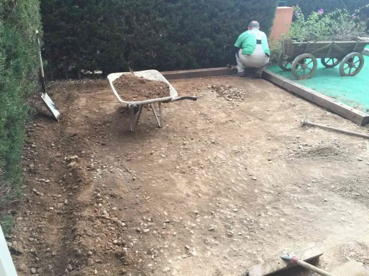 Una persona esparce tierra en un patio trasero con una carretilla y herramientas de jardinería.