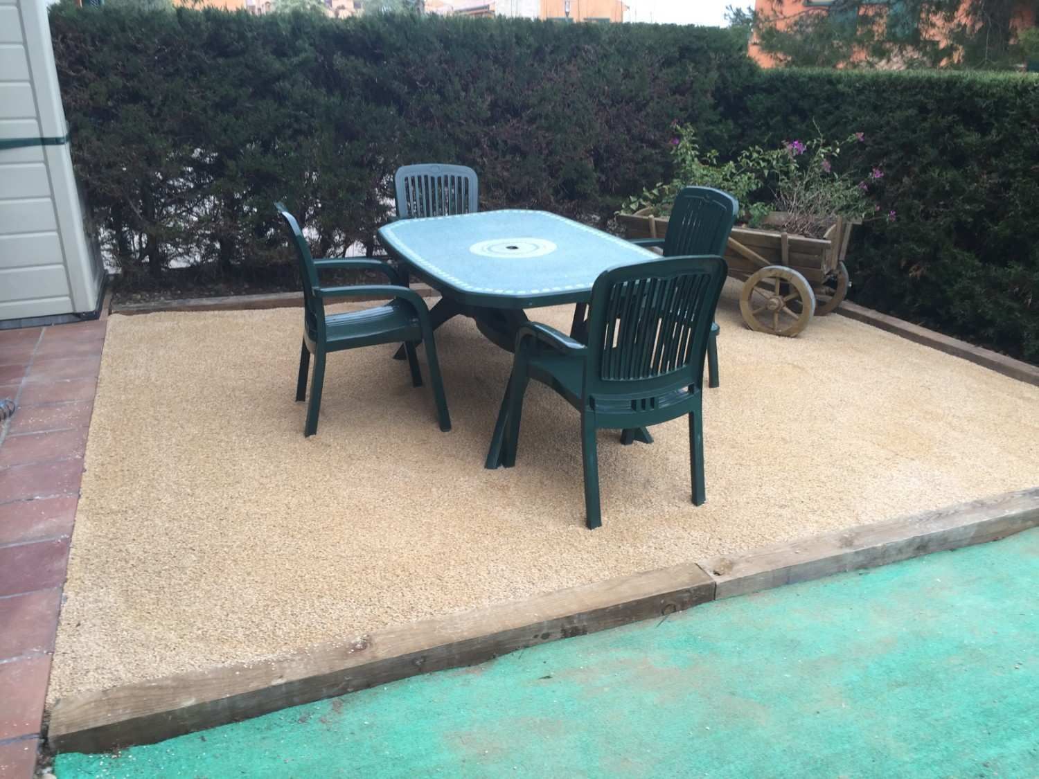 Patio con mesa redonda azul y cuatro sillas oscuras junto a un seto y el borde de la piscina de color turquesa.