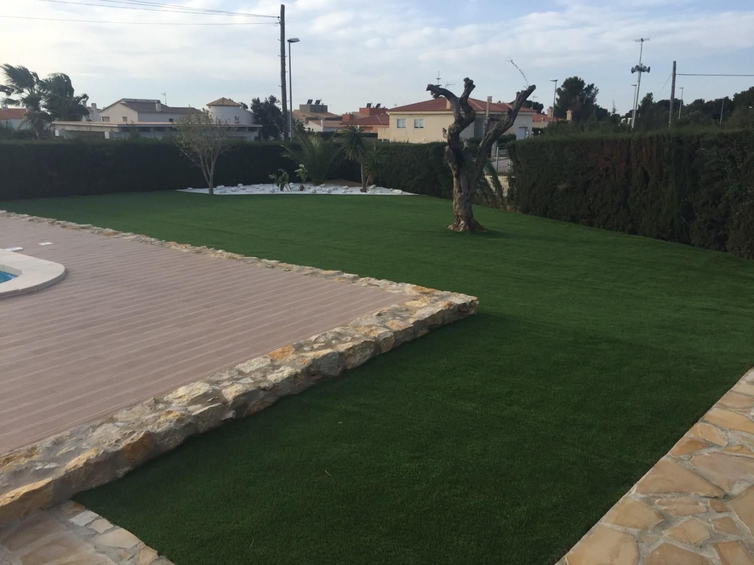 Patio trasero con césped artificial, bordes de piedra y un árbol podado cerca de los setos.