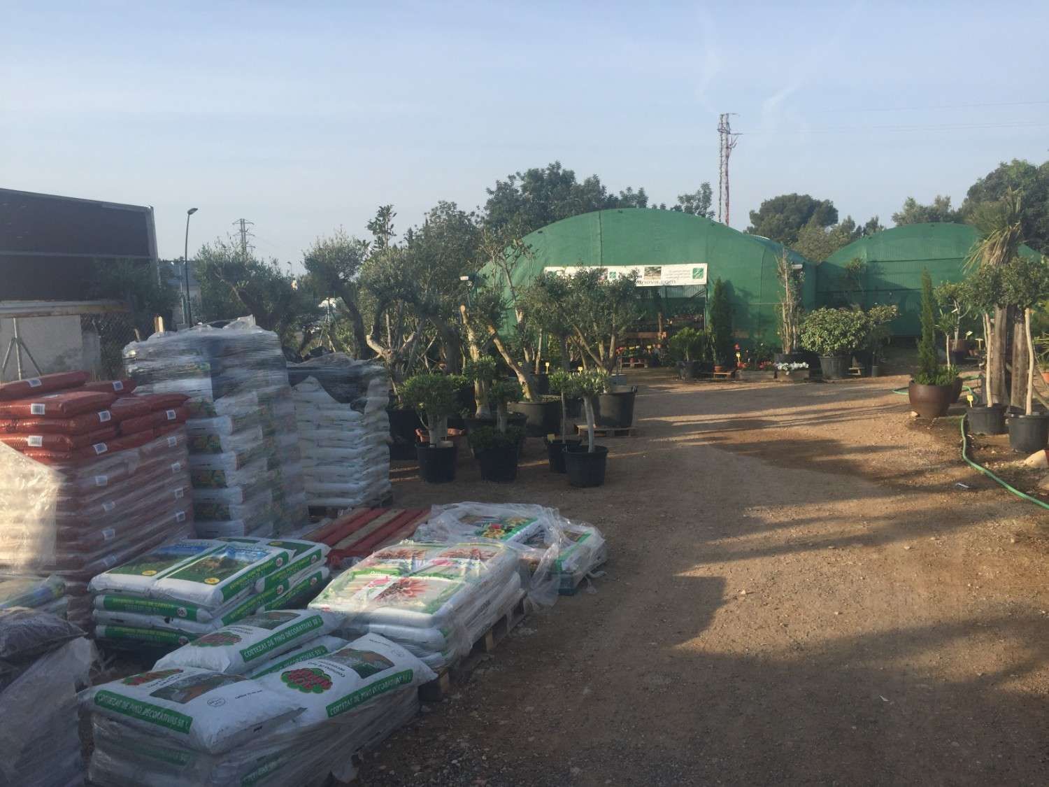 Centro de jardinería con sacos de tierra apilados, plantas en macetas y toldos verdes a lo largo de un camino de tierra.