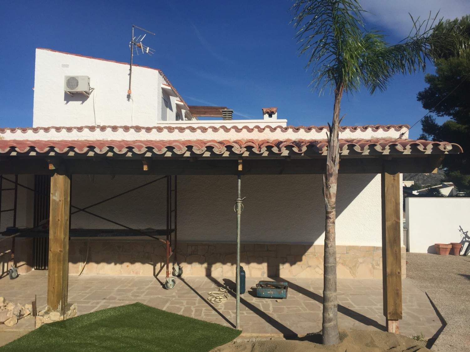 Patio trasero con techo de tejas, palmeras y zona de estar a la sombra bajo un cielo azul brillante.