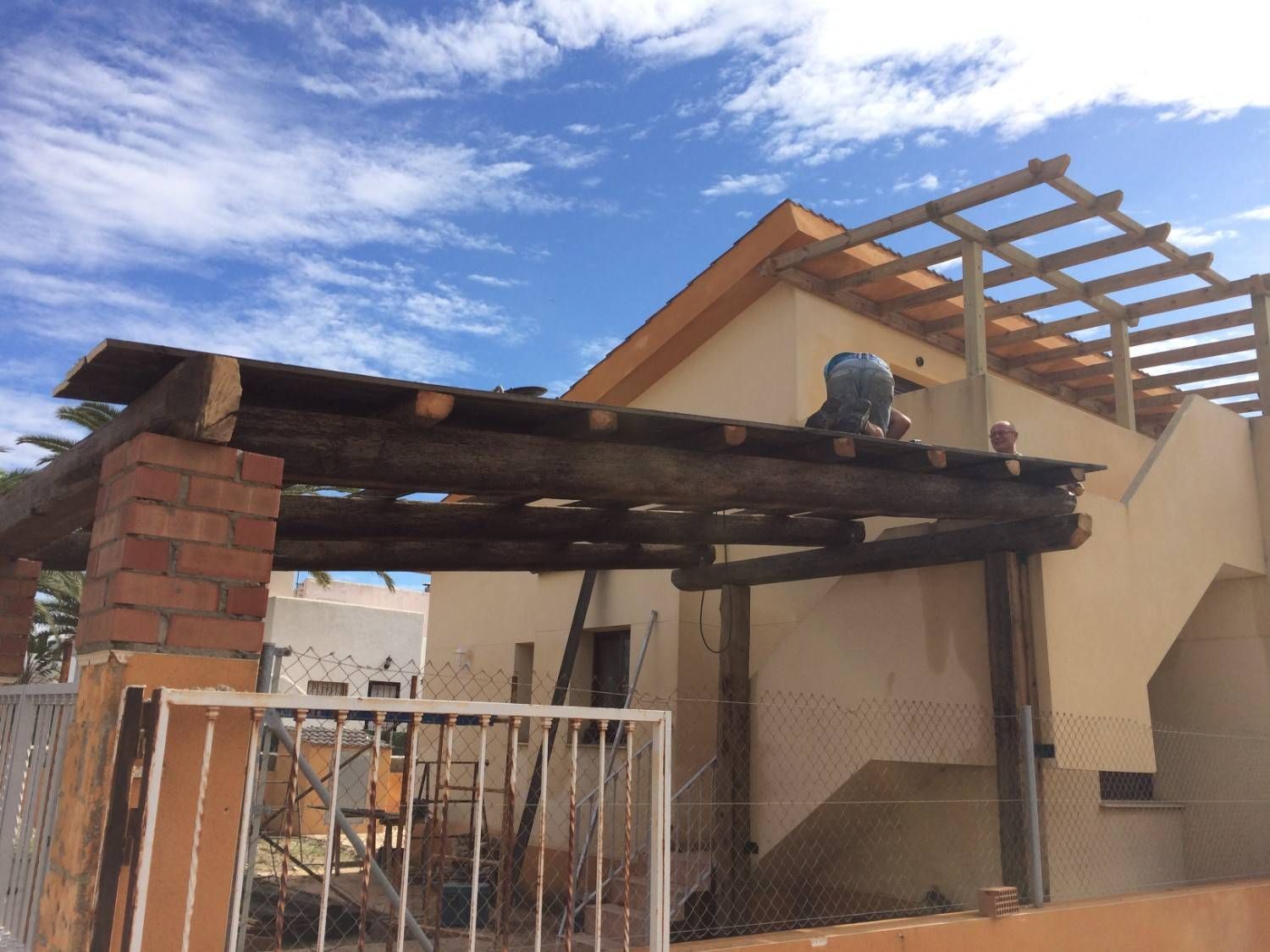 Cochera de madera dañada, adosada a una casa, con vigas a la vista y una puerta de metal bajo un cielo azul.