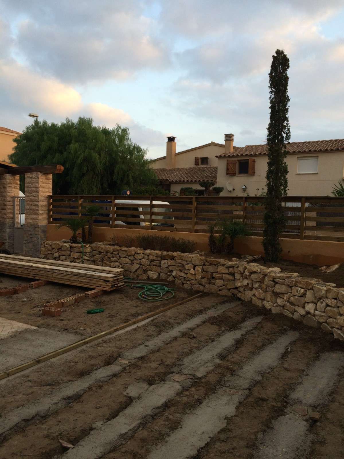 Patio de piedra y jardín trasero ajardinado con valla de madera, muro de contención y un alto ciprés al atardecer.