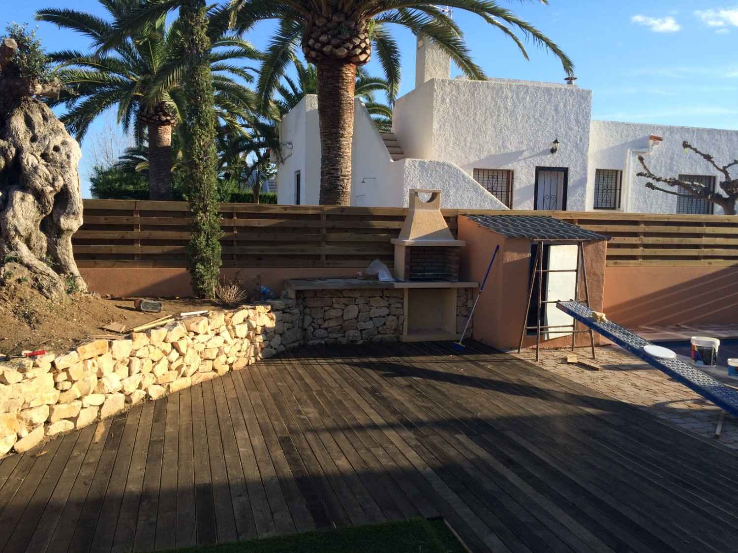Patio soleado con palmeras, terraza de madera, muros de piedra y tumbonas junto a la piscina.