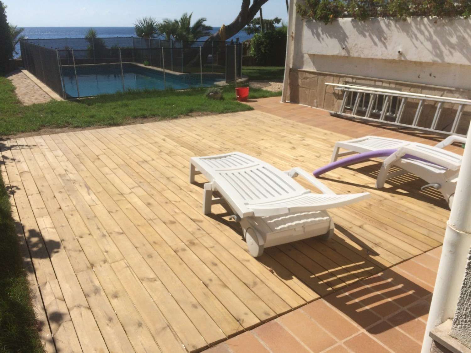 Patio de madera soleado con tumbonas, piscina y vistas al océano más allá de un césped.