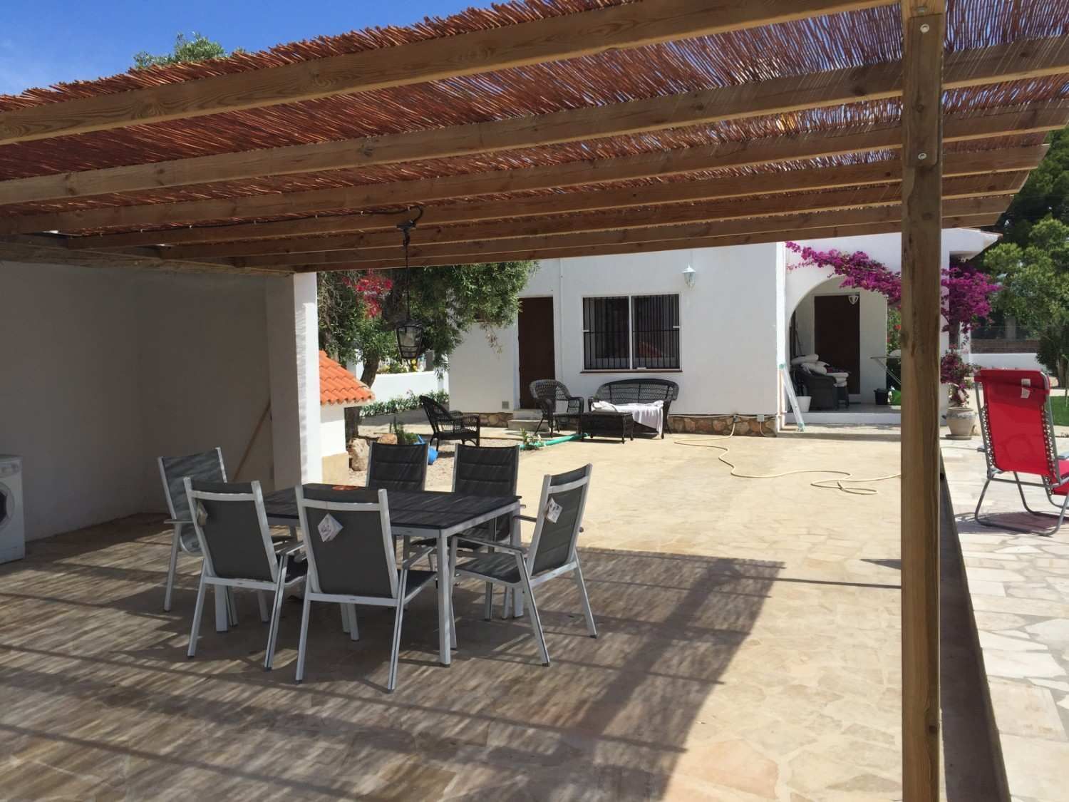 Patio con pérgola de madera, mesa de comedor exterior, sillas y una casa blanca al fondo.