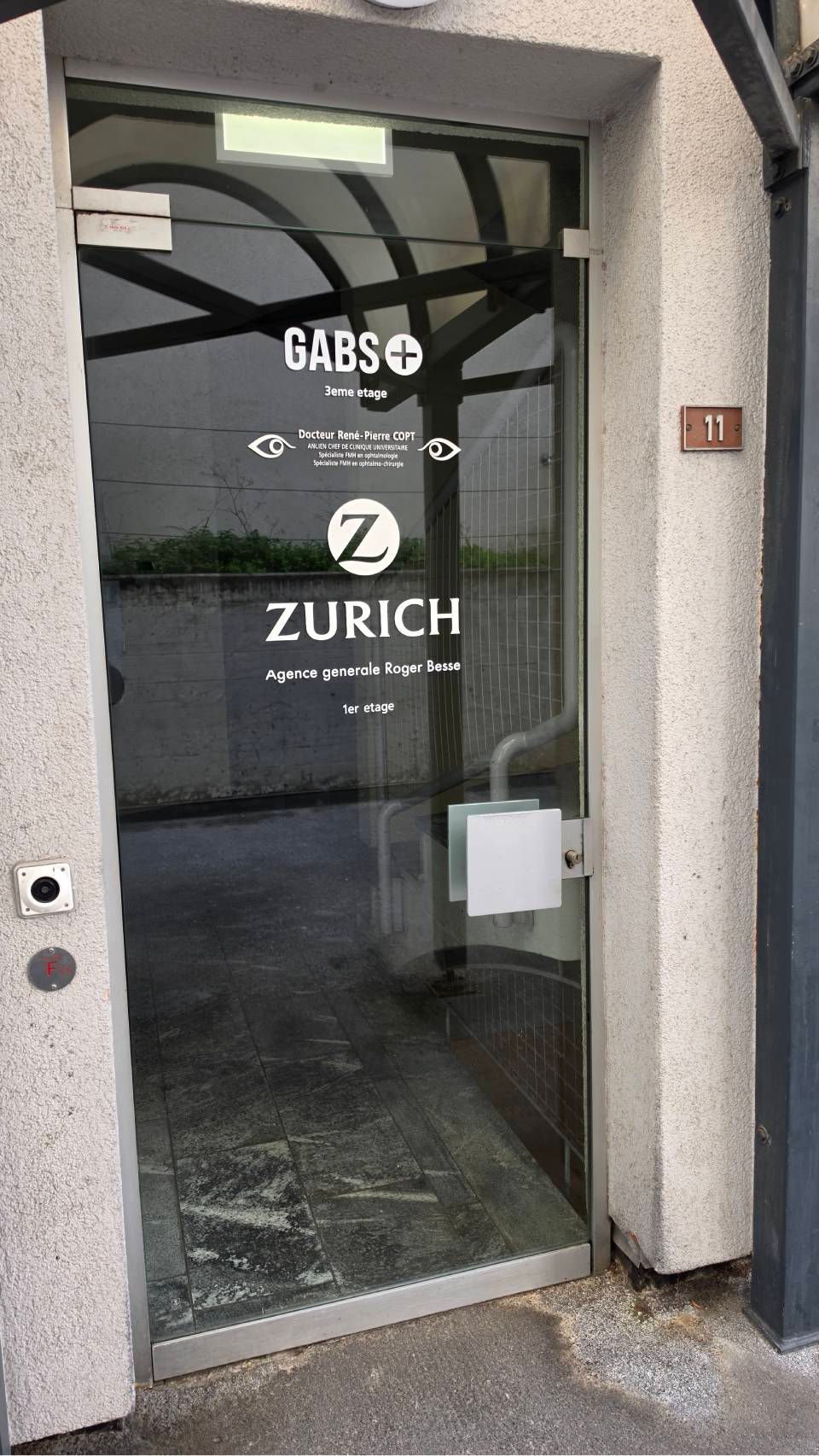 Porte d'entrée vitrée avec logo Zurich et signalétique blanche sur un mur gris dans le hall d'entrée d'un bâtiment.