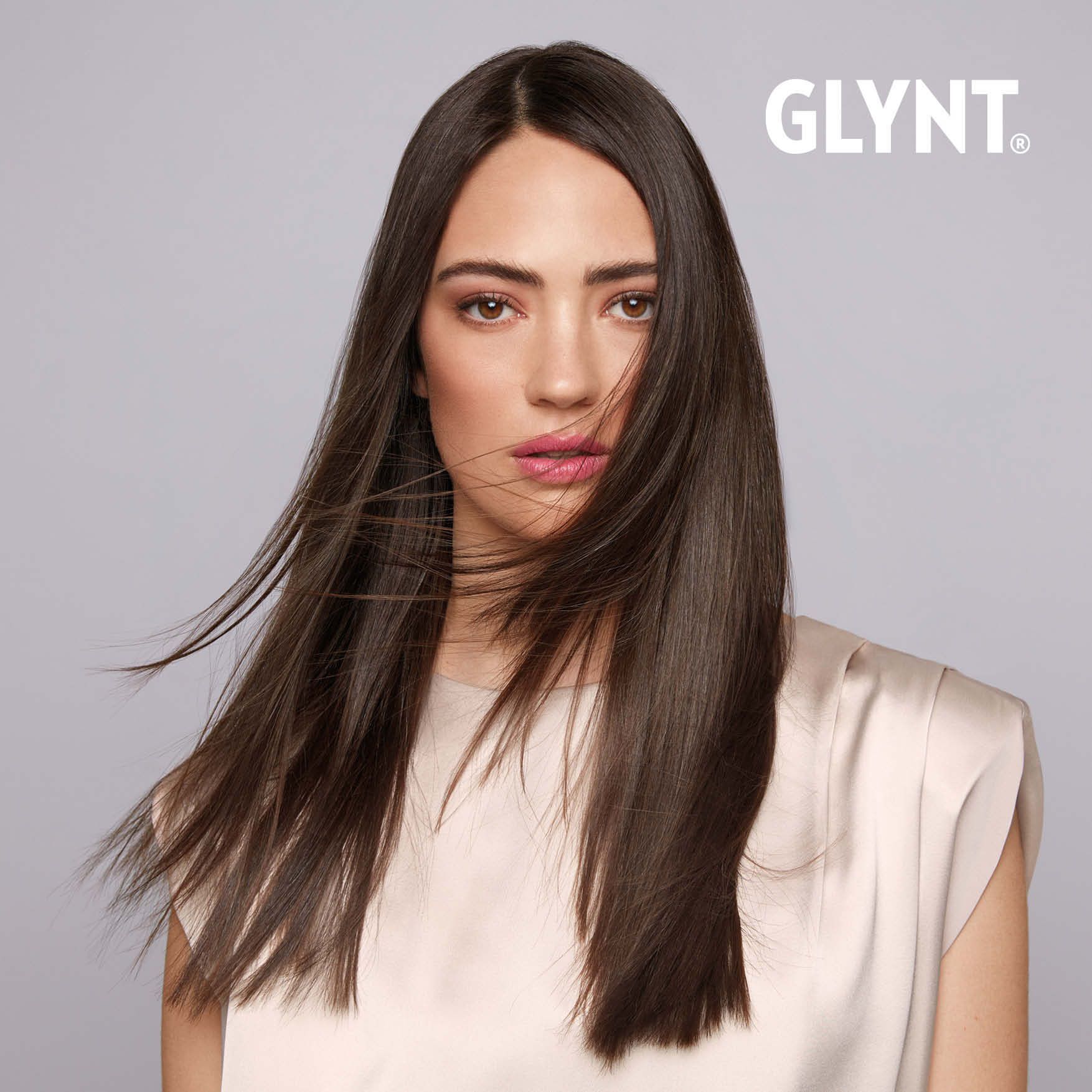 Frau mit langem, glattem, dunklem Haar; neutraler Hintergrund; trägt ein helles Oberteil; das Haar weht ihr ins Gesicht.