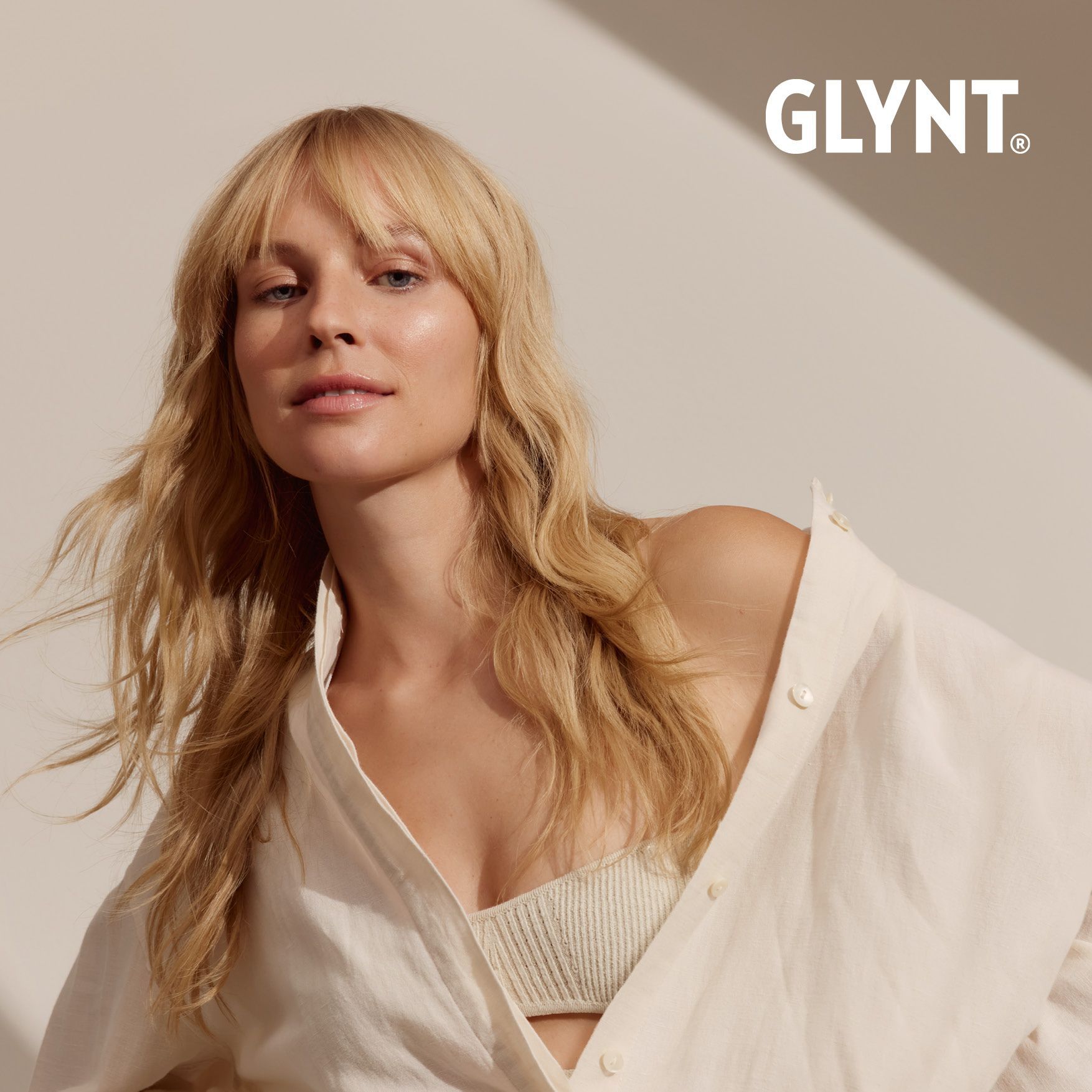 Frau mit blonden Haaren und weißem Hemd, blickt in die Kamera. GLYNT-Logo in der Ecke.