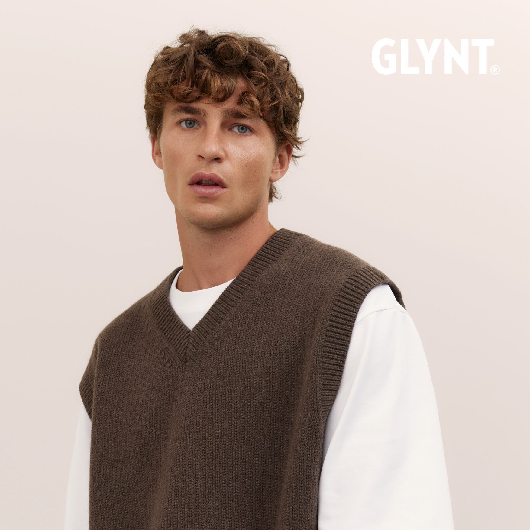 Mann in brauner Weste über weißem Hemd, neutraler Hintergrund.