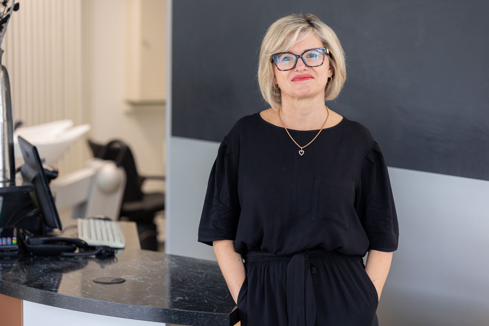 Frau mit blonden Locken, Brille, lächelnd, mit schwarzem Oberteil, steht vor einem Fenster.
