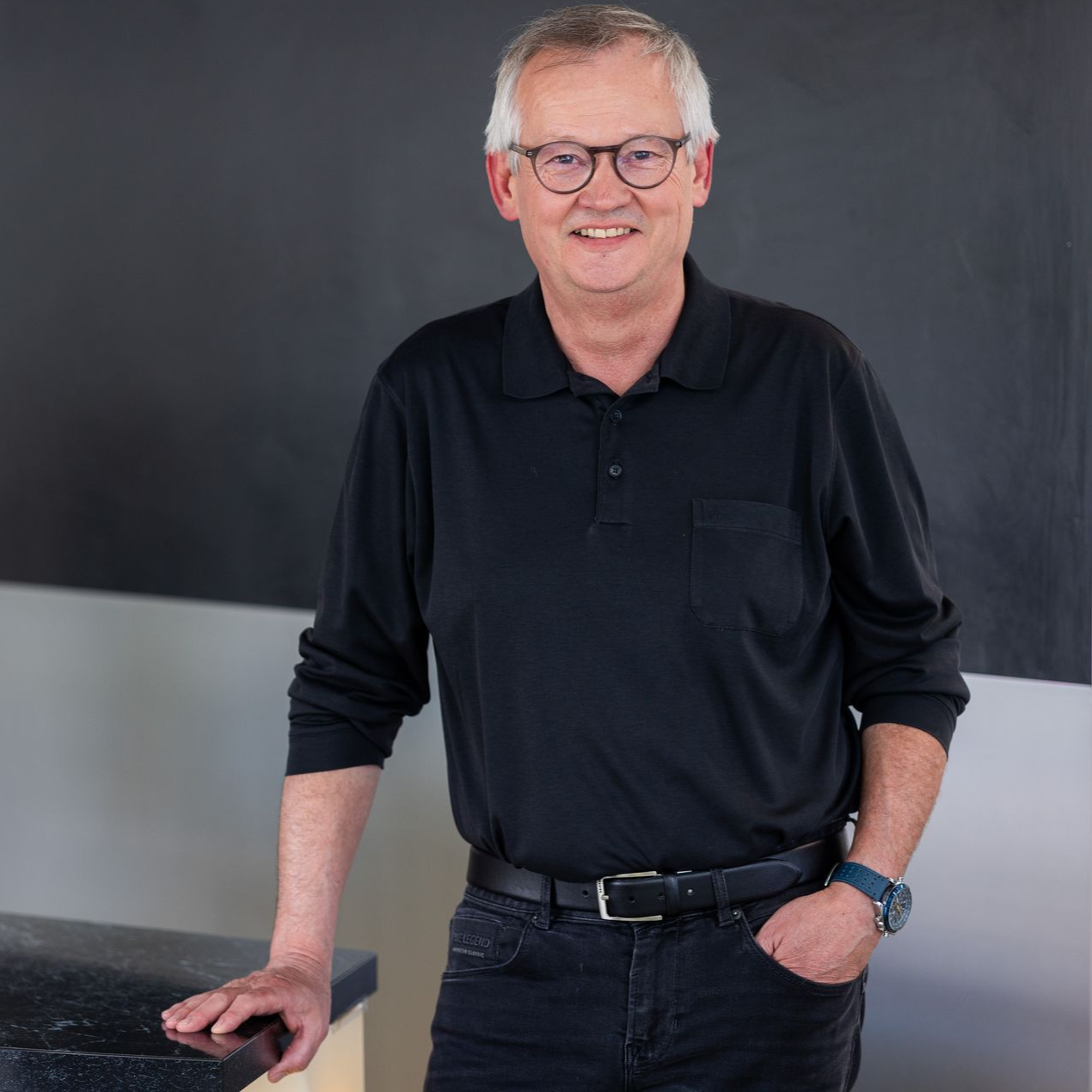 Lächelnder Mann mit Brille, kurzen, ergrauenden Haaren und schwarzem Hemd vor einem verschwommenen blauen Hintergrund.