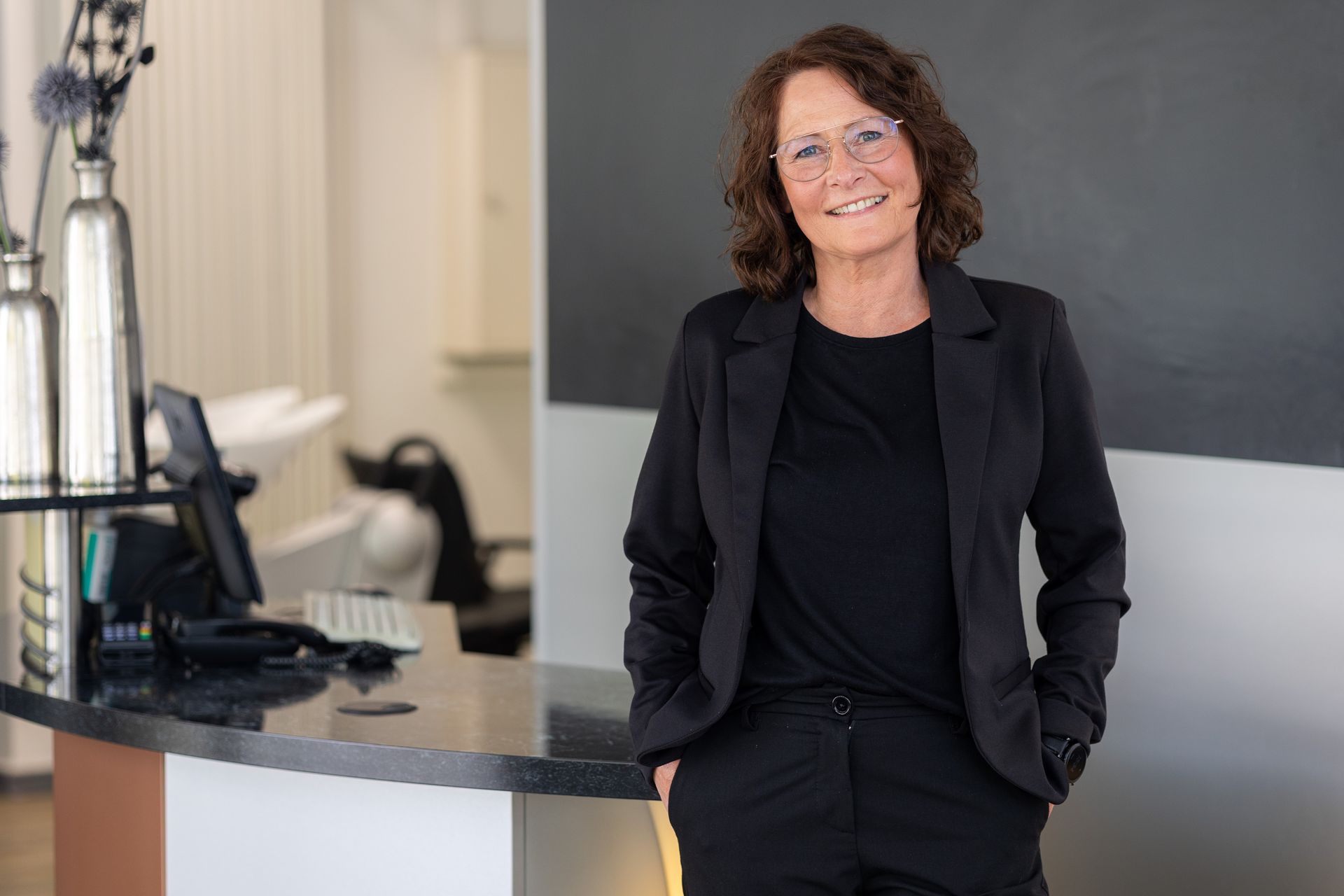 Lächelnde Frau mit kurzen braunen Haaren, schwarzem Oberteil, vor blauem Hintergrund.