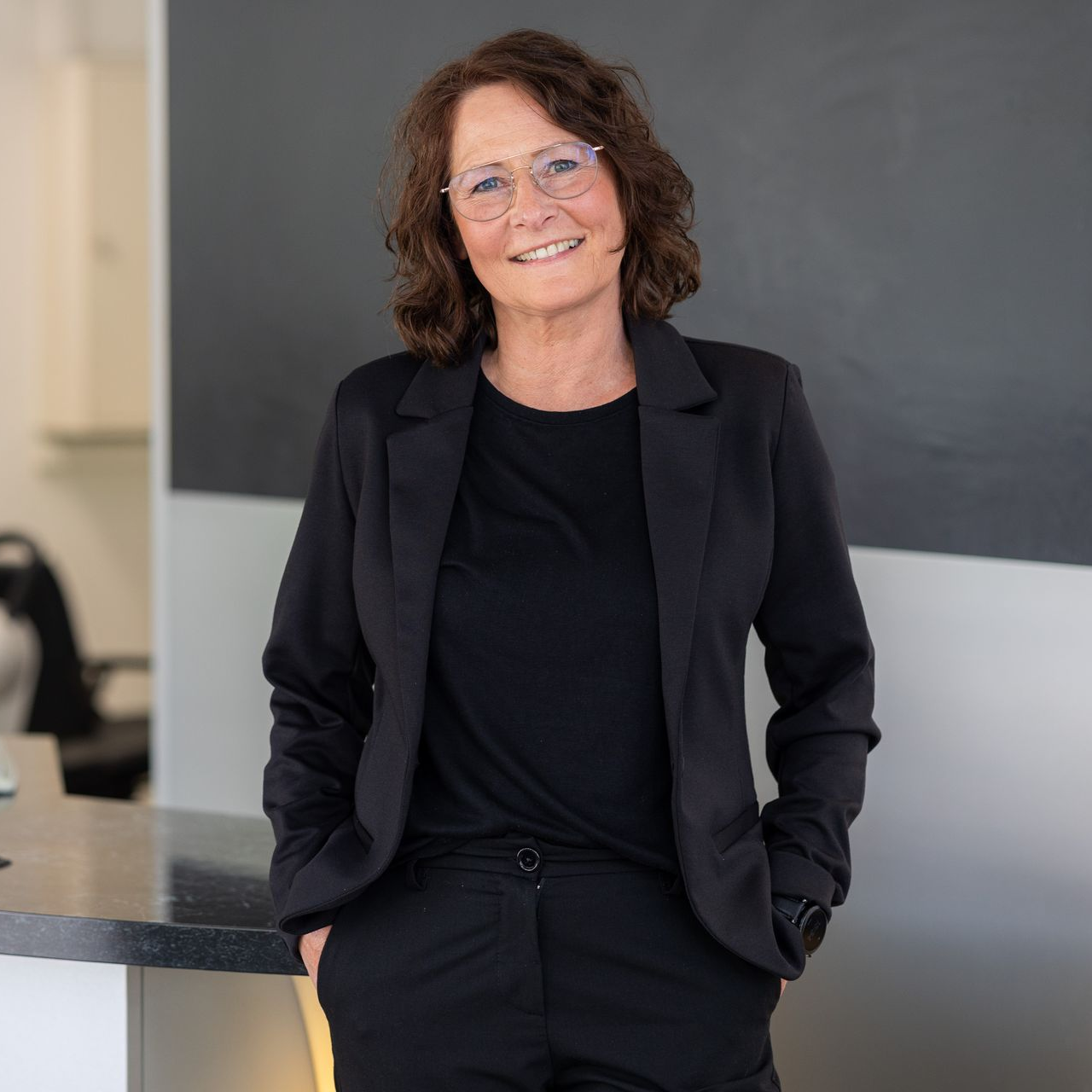 Lächelnder Mann mit Brille, kurzen, ergrauenden Haaren und schwarzem Hemd vor einem verschwommenen blauen Hintergrund.