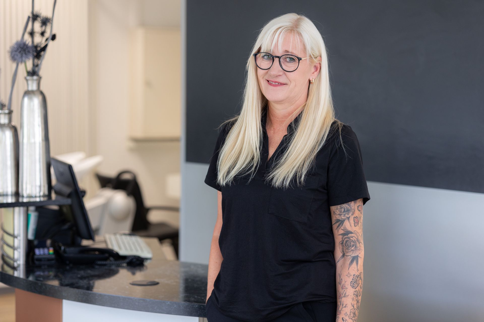 Frau mit blonden Haaren und Brille, lächelnd, trägt ein schwarzes Oberteil vor einem hellblauen Hintergrund.