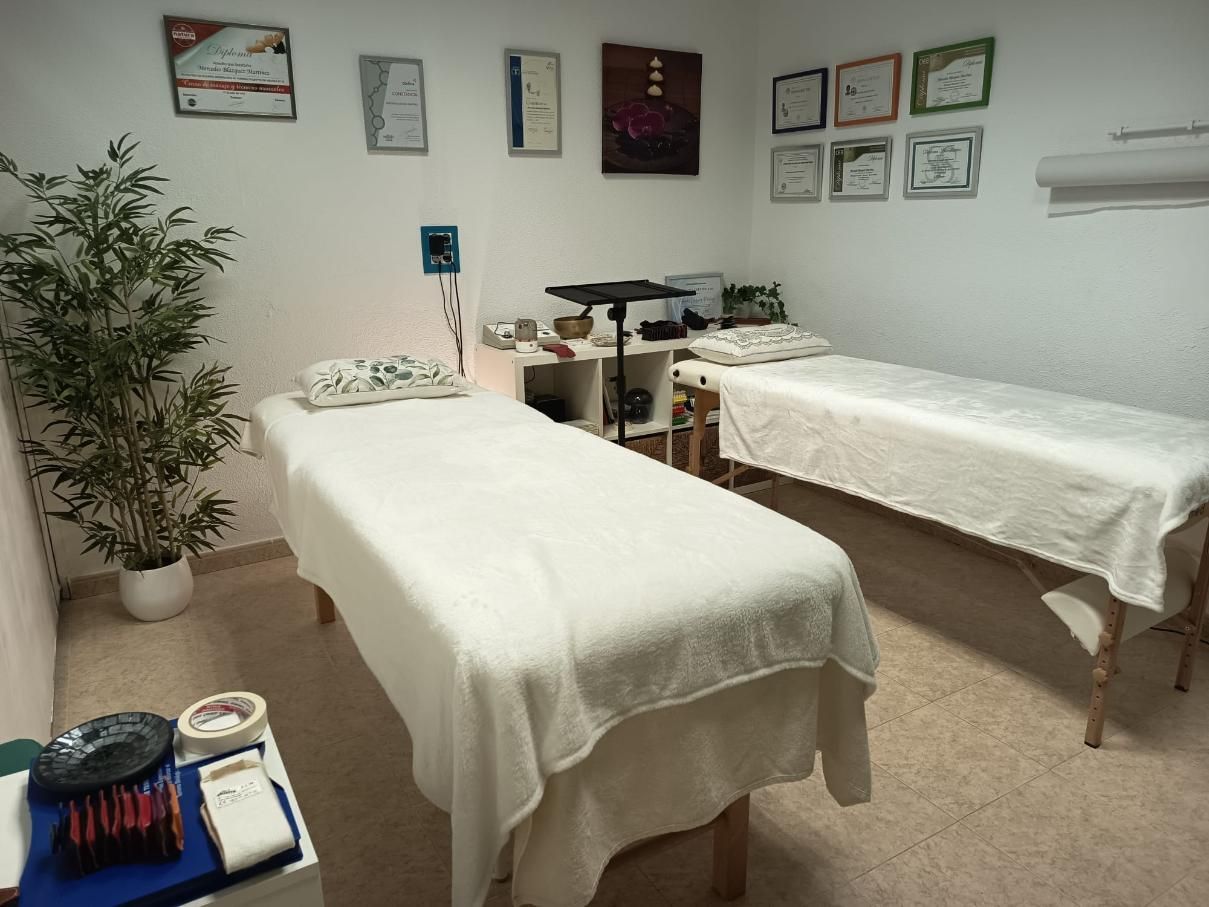 Sala de masajes con dos camillas de tratamiento, sábanas blancas, cuadros en las paredes y una planta en maceta.