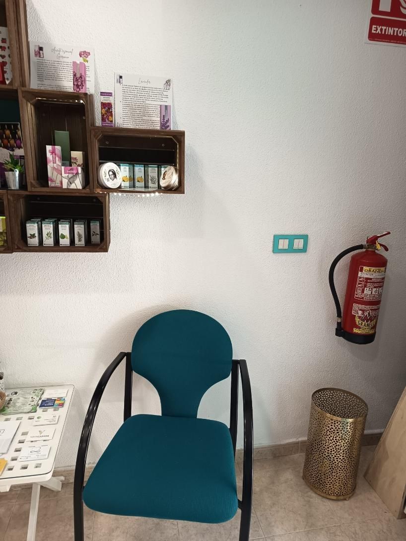 Zona de espera con silla color turquesa, estantes en la pared, botellas de desinfectante y un extintor de incendios.