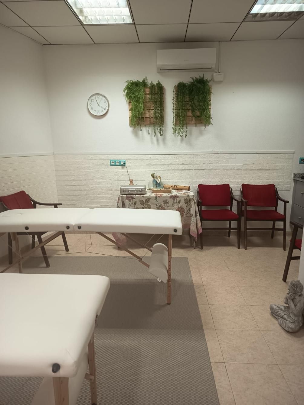 Pequeña sala de espera de una clínica con camilla blanca, sillas rojas, reloj de pared y plantas colgantes.