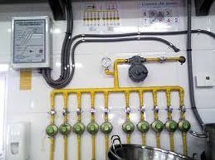 Colector de gas montado en la pared con tuberías amarillas y múltiples cilindros verdes en un cuarto de servicio.