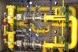 Tuberías y válvulas industriales amarillas en un sistema mecánico, con manómetros verdes sobre un fondo gris.