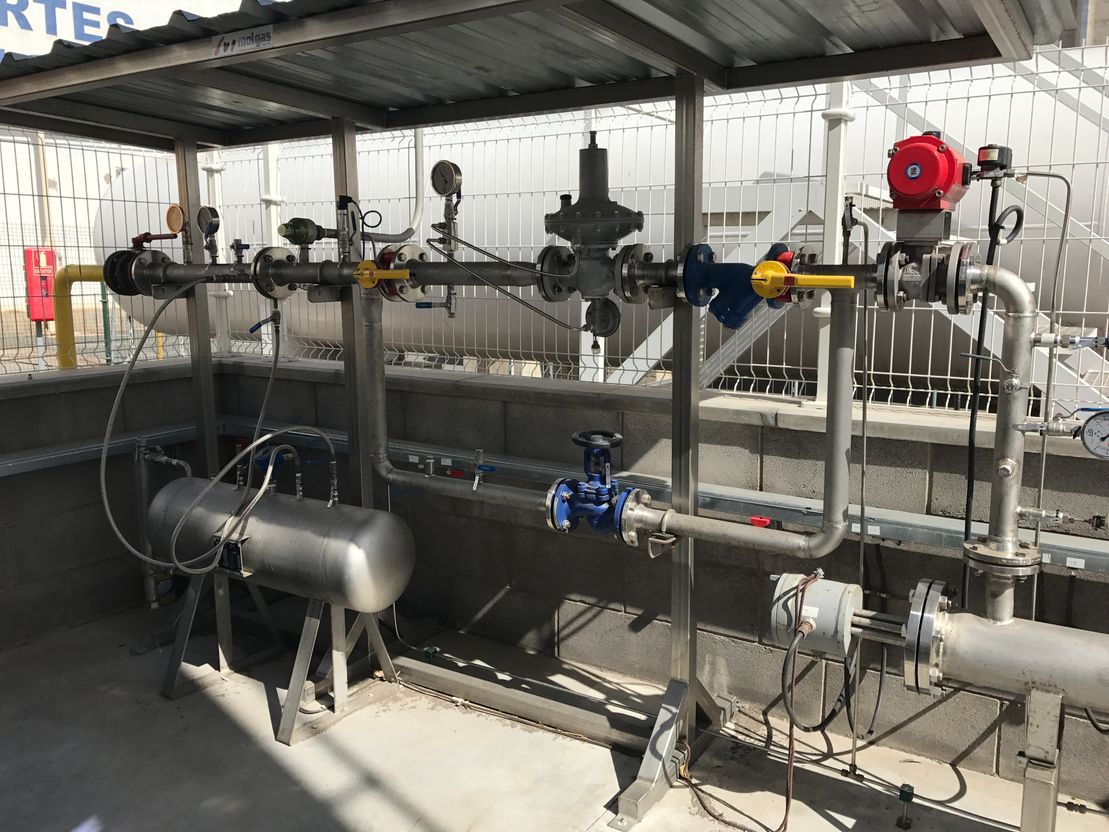 Tuberías y válvulas industriales dentro de un área de maquinaria cubierta, con un tanque plateado y equipos de control.