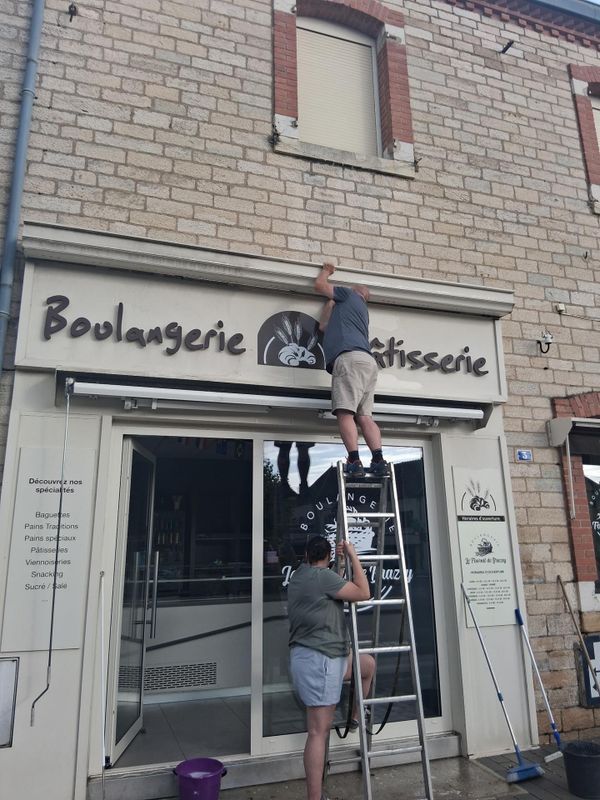 Deux personnes changent une enseigne devant une boulangerie, l'une sur une échelle et l'autre debout en dessous.