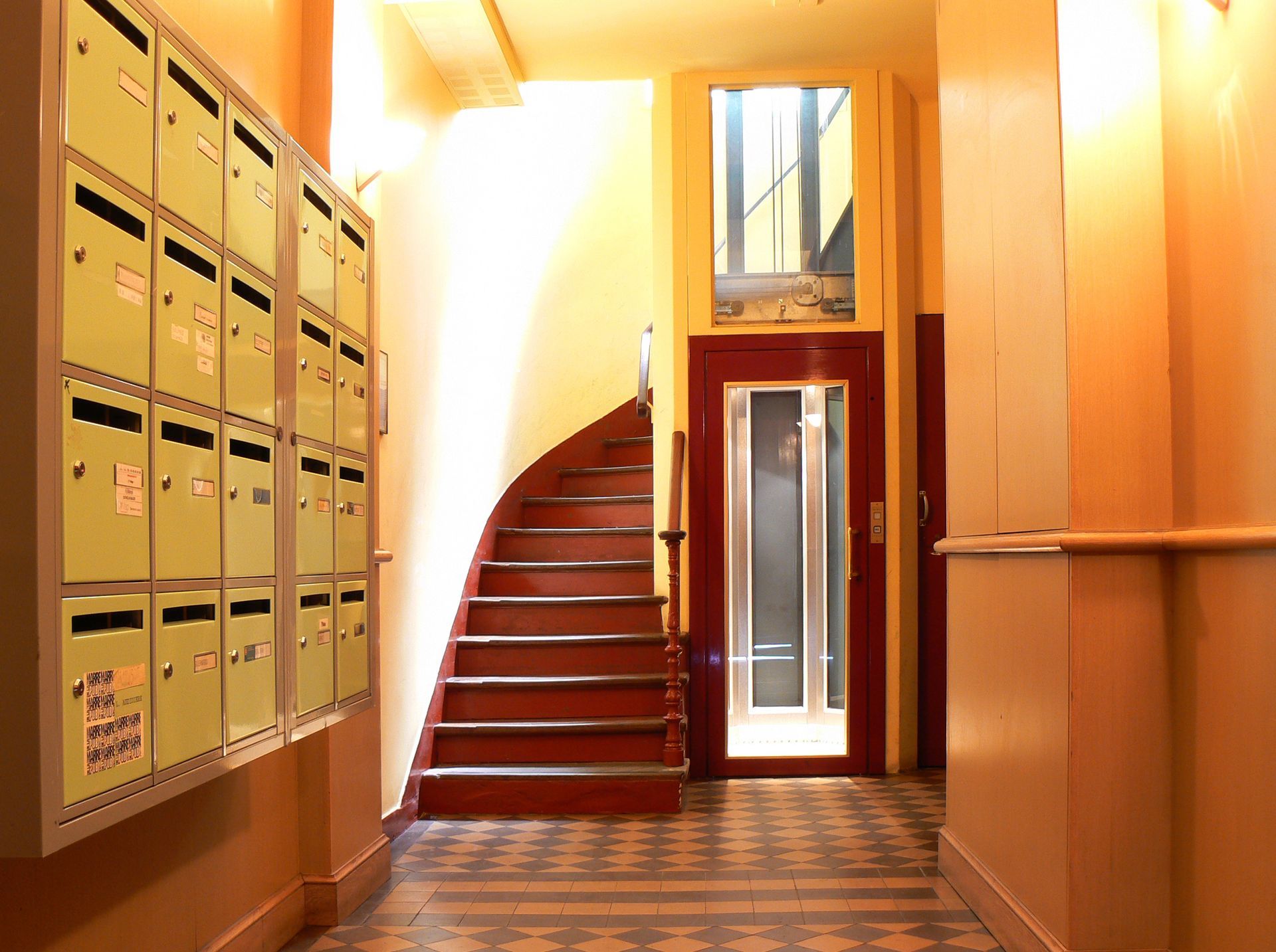 Couloir avec boîtes aux lettres à gauche, escalier rouge incurvé et éclairage jaune chaud.