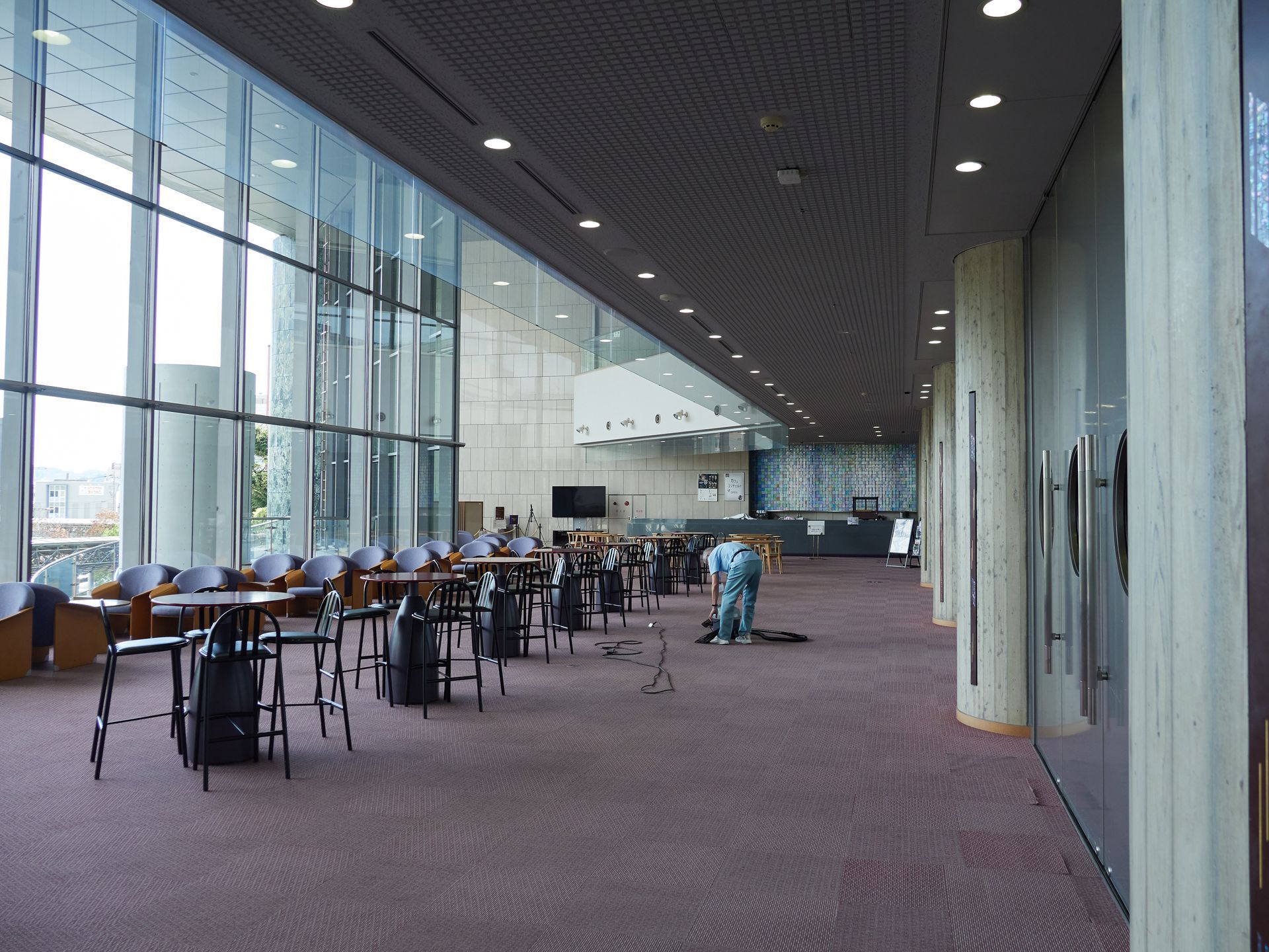 Un atrium moderne et spacieux avec de hautes parois vitrées, des tables et quelques personnes qui marchent sur un sol recouvert de moquette.