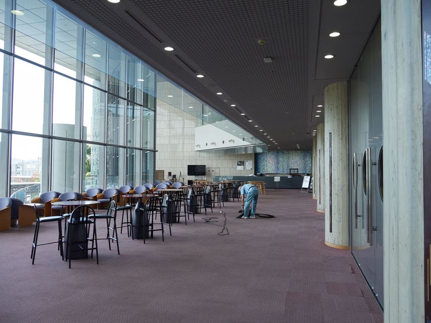 Hall d'entrée moderne et spacieux avec tables, chaises, hautes baies vitrées et quelques personnes à l'intérieur.