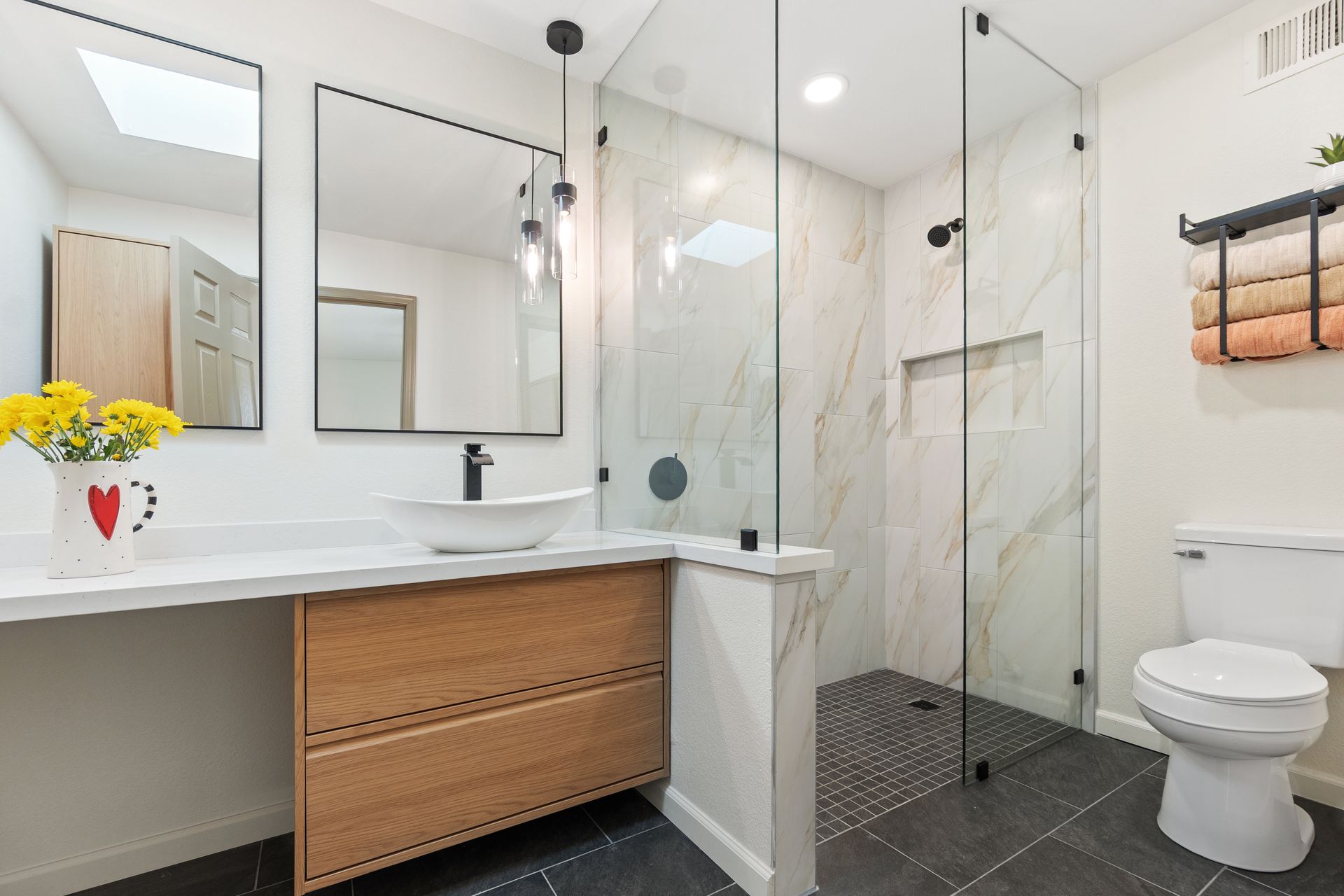 Salle de bains moderne avec double vasque, douche vitrée et toilettes sur carrelage foncé.