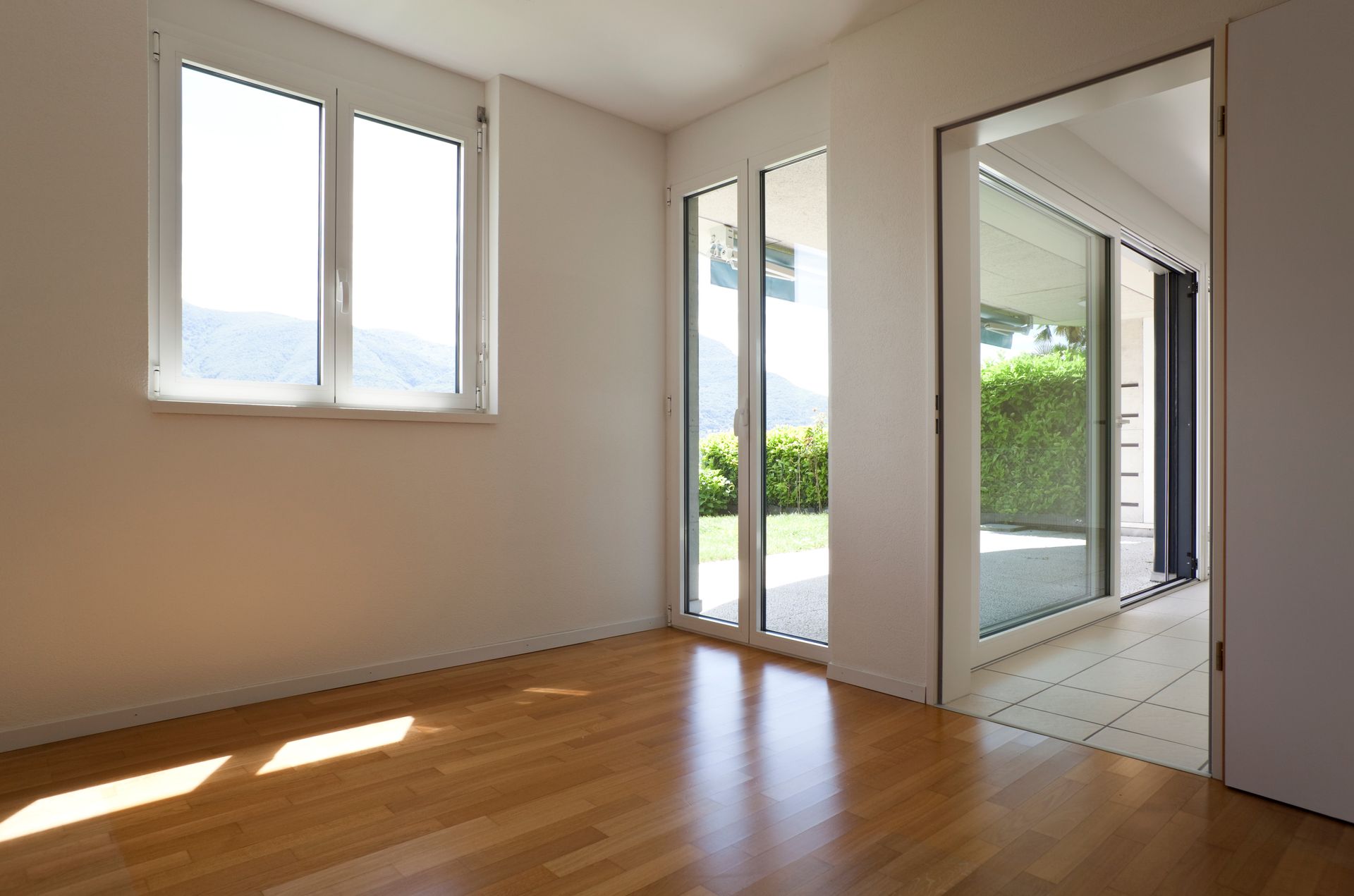 Pièce vide et ensoleillée avec parquet, murs blancs et grandes fenêtres donnant sur un patio et de la verdure.