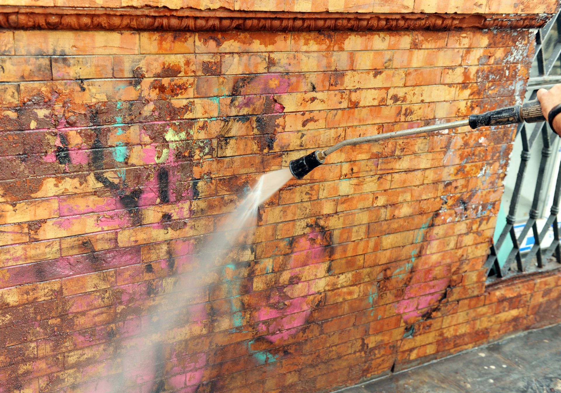 Un nettoyeur haute pression enlève des graffitis d'un mur de briques près d'une grille de fenêtre.