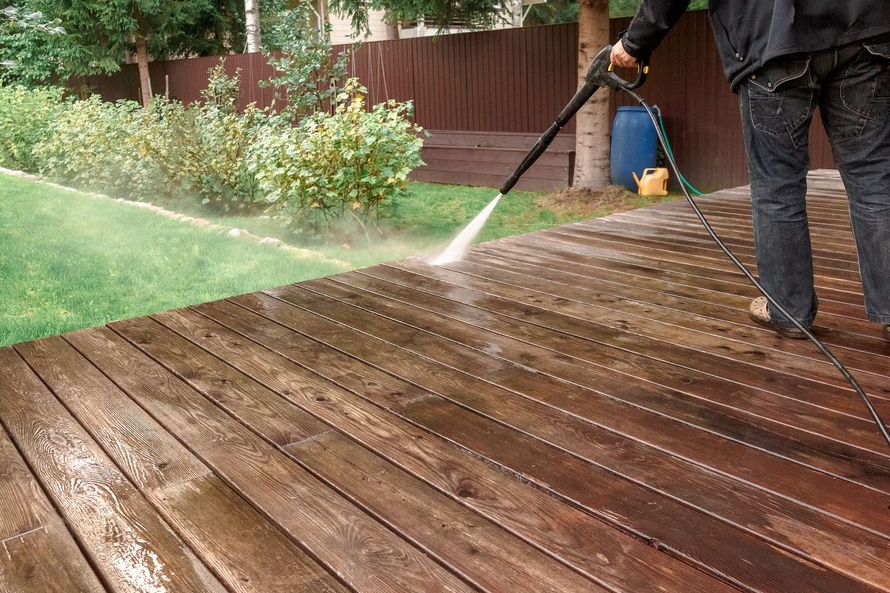 Une personne nettoie à haute pression une terrasse en bois avec un tuyau d'arrosage dans un jardin.