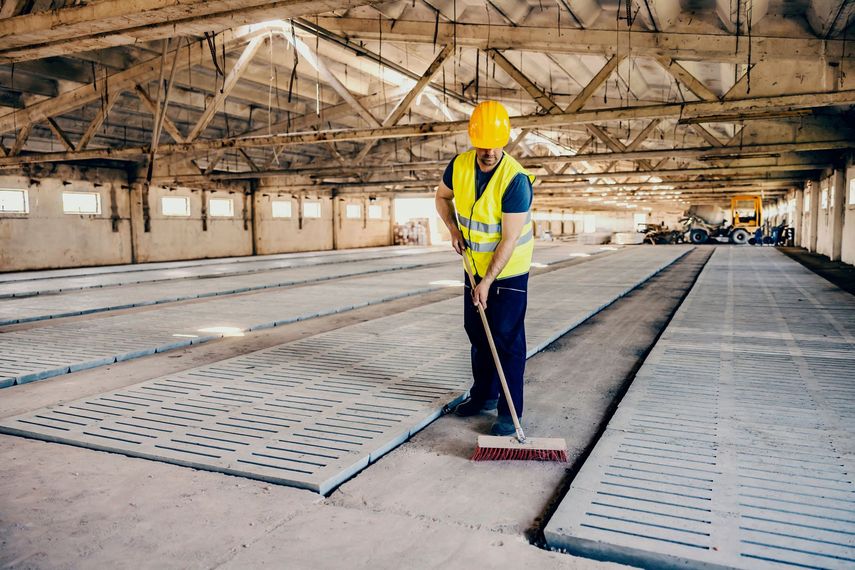 Ouvrier portant un casque jaune balayant le sol poussiéreux d'un entrepôt industriel.