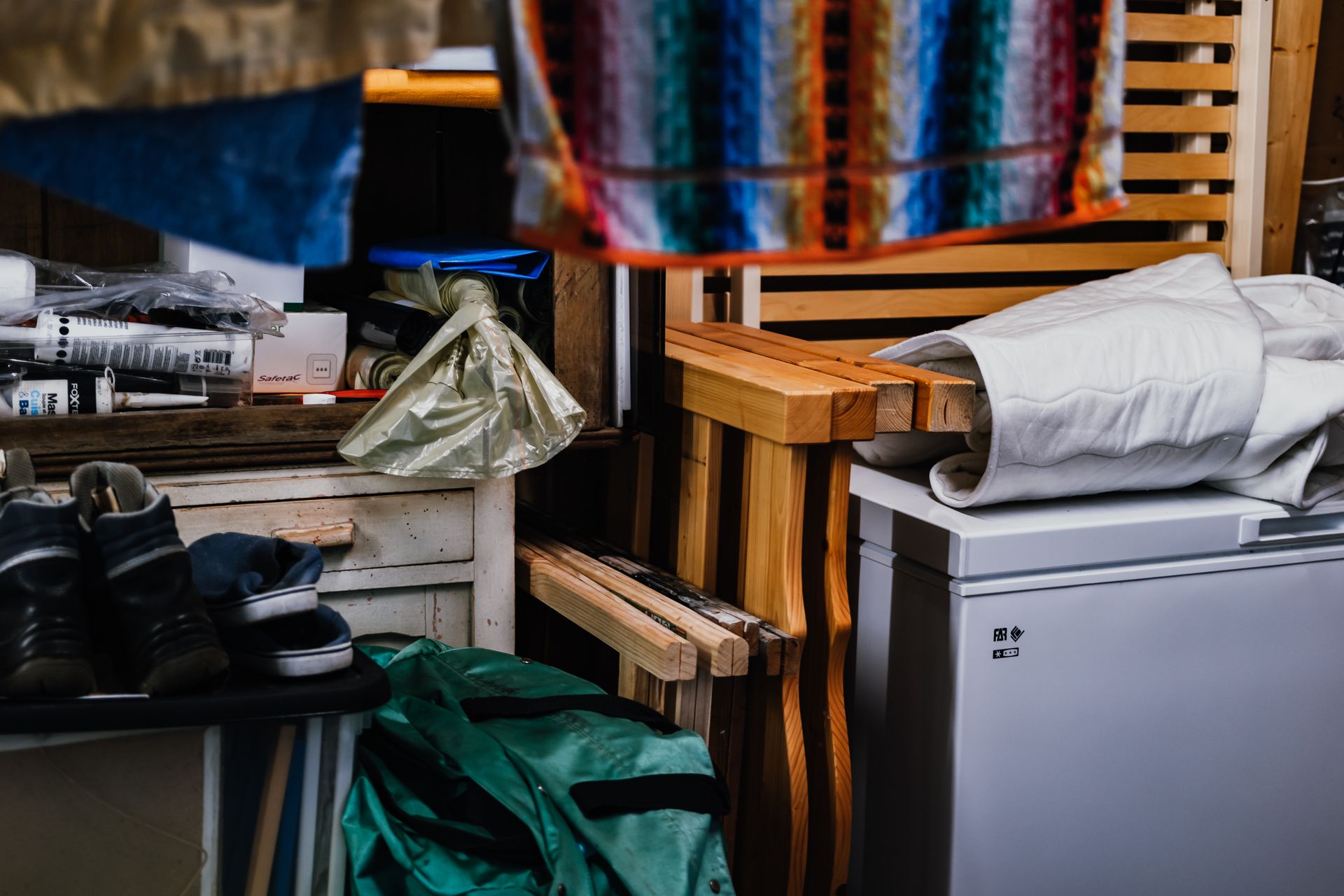 Espace de rangement encombré avec des caisses en bois empilées, un minifrigo, des chaussures et des tissus colorés suspendus.