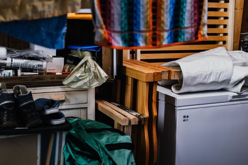 Espace de rangement encombré avec des caisses en bois empilées, un minifrigo, des chaussures et des tissus colorés suspendus.