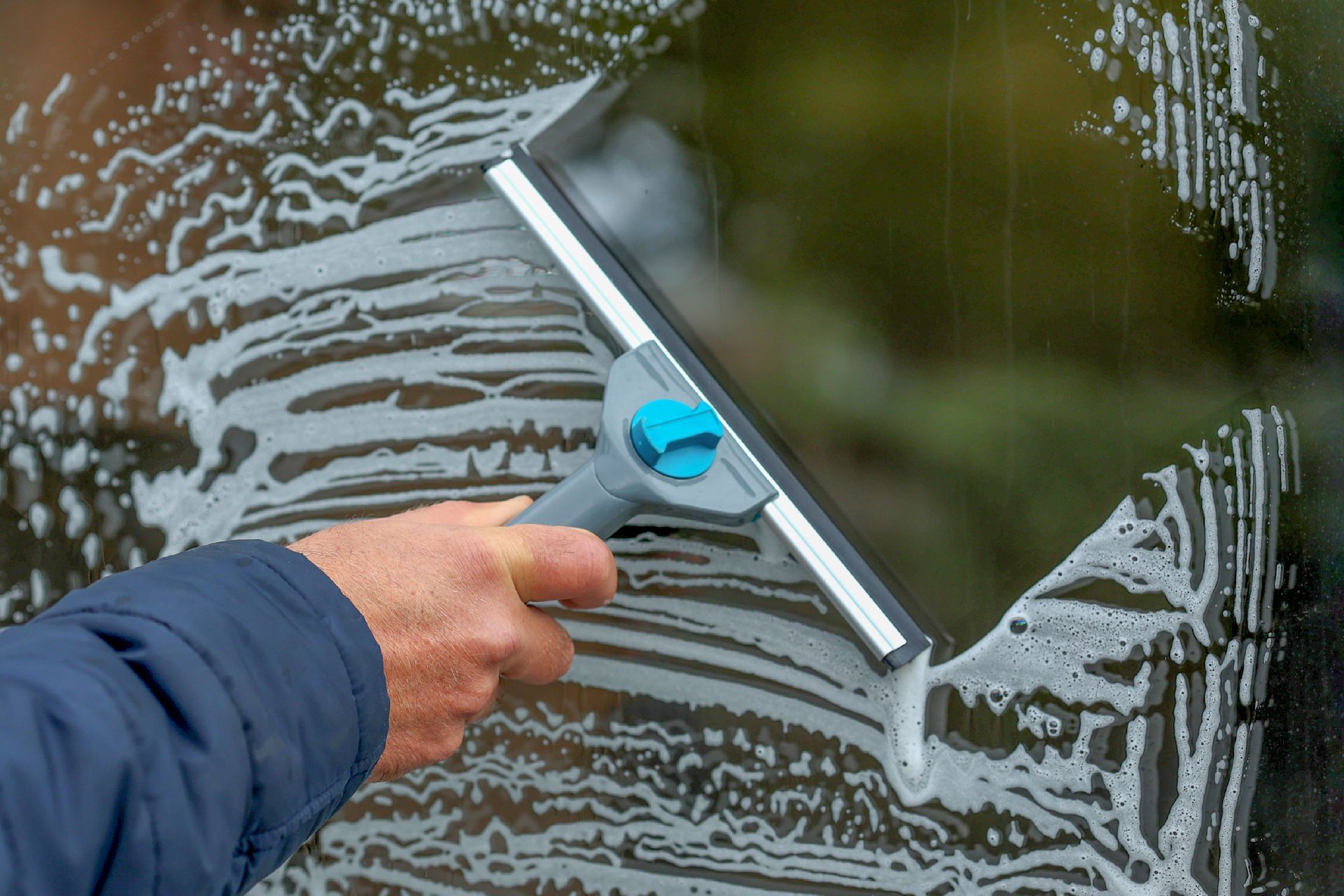 Une personne raclant de l'eau savonneuse sur une vitre de voiture avec un outil à manche bleu.