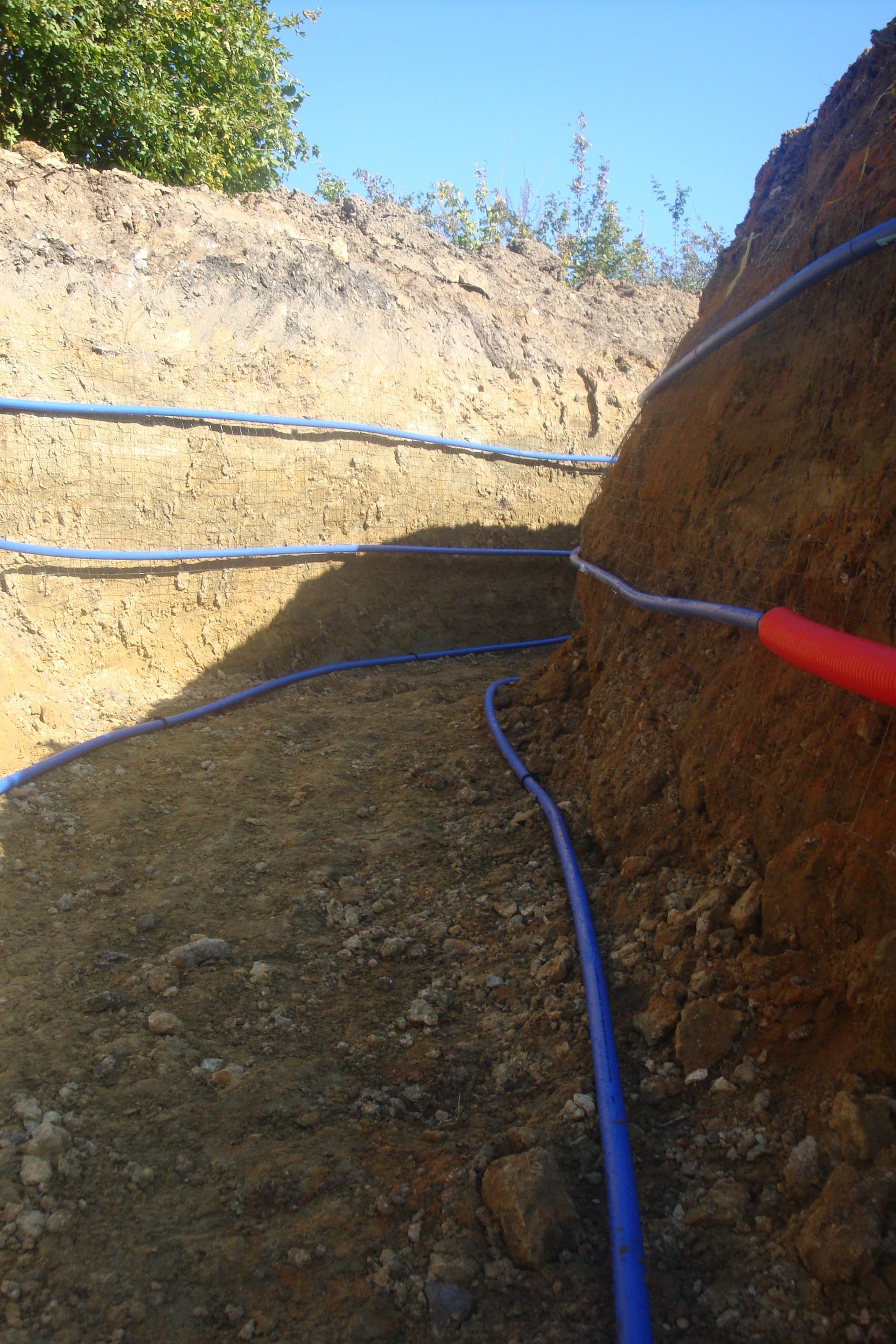 Tranchée creusée dans la terre, avec des tuyaux bleus et un fil rouge.