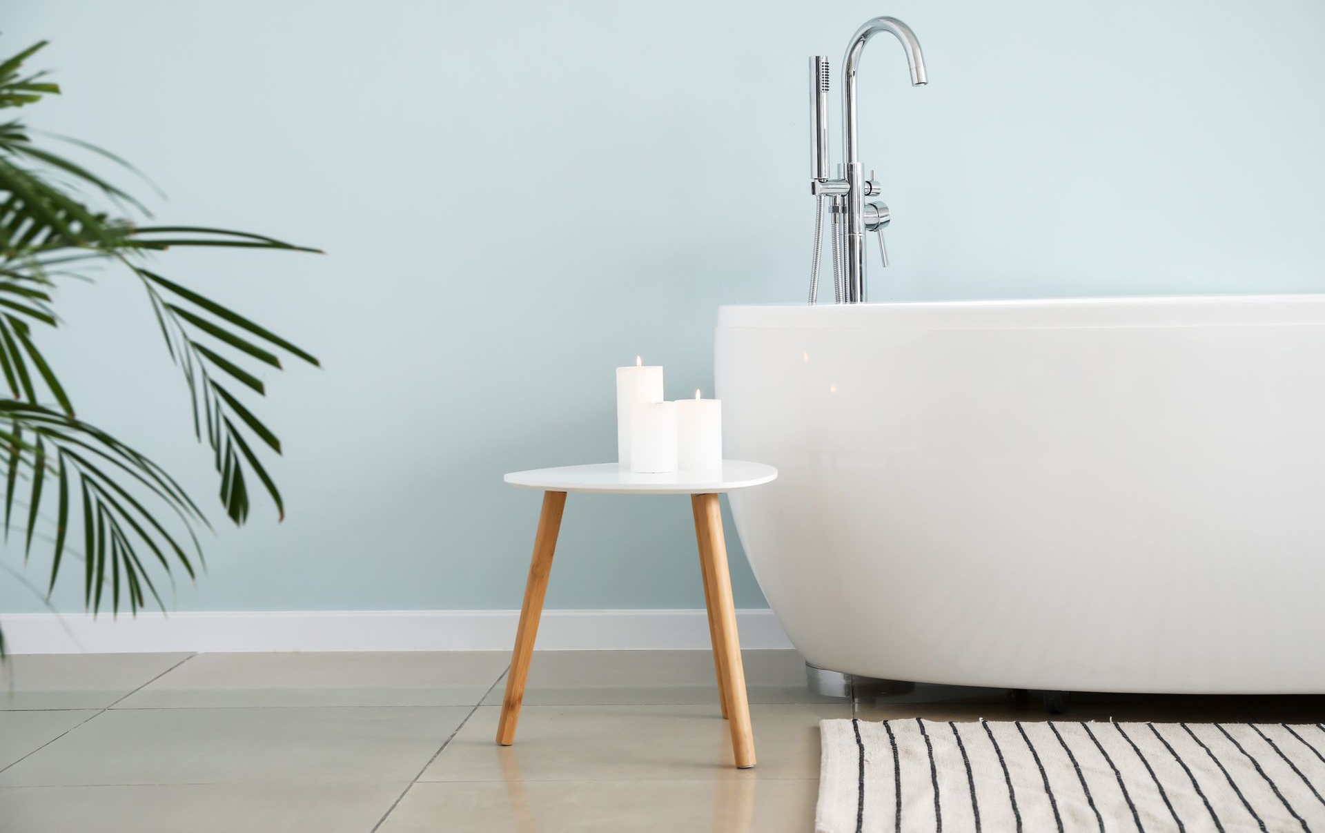 Baignoire blanche, table avec bougies, palmier, mur bleu clair, tapis noir et blanc.