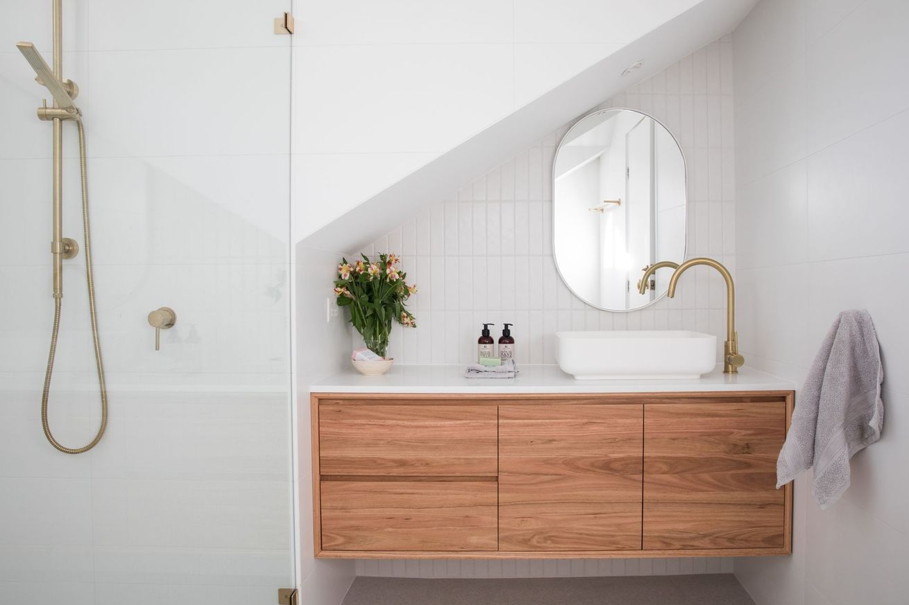 Une salle de bains avec meuble en bois, miroir ovale, lavabo blanc et robinetterie dorée.