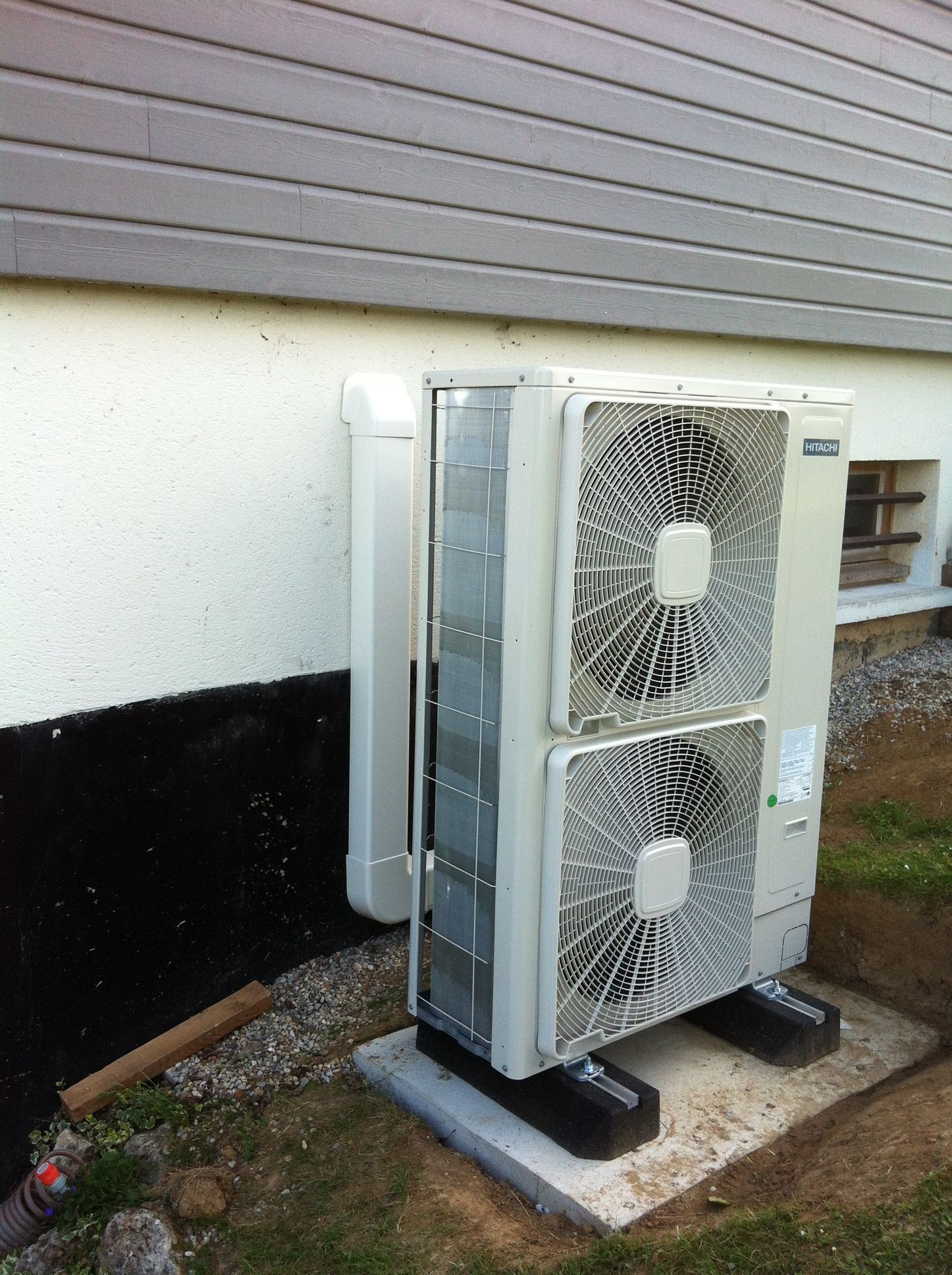 Un climatiseur, de couleur beige clair, est fixé sur du béton près d'un bâtiment blanc au revêtement gris.