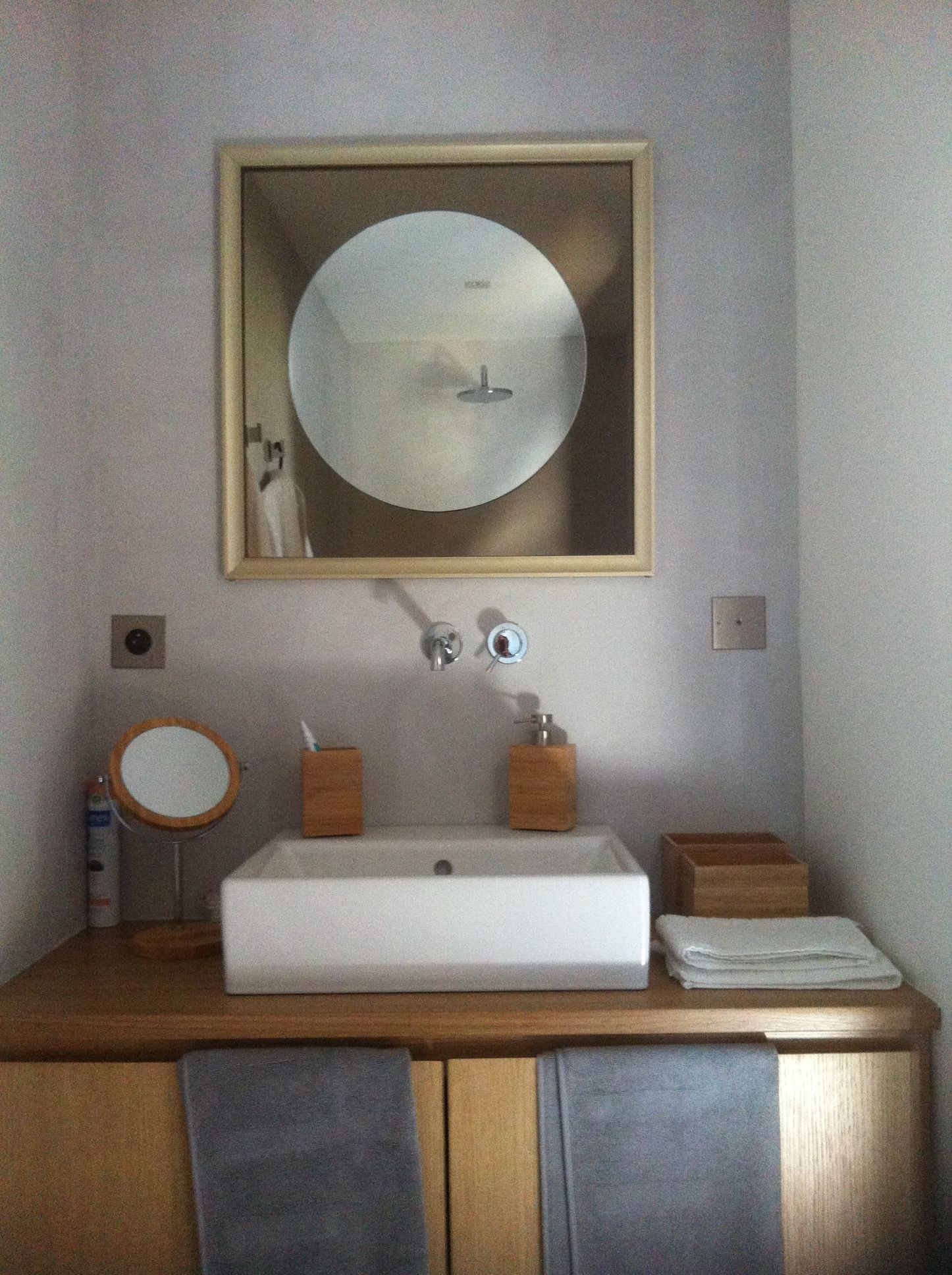 Meuble avec vasque carrée, miroir rond et armoire en bois. Des serviettes grises sont suspendues en dessous.