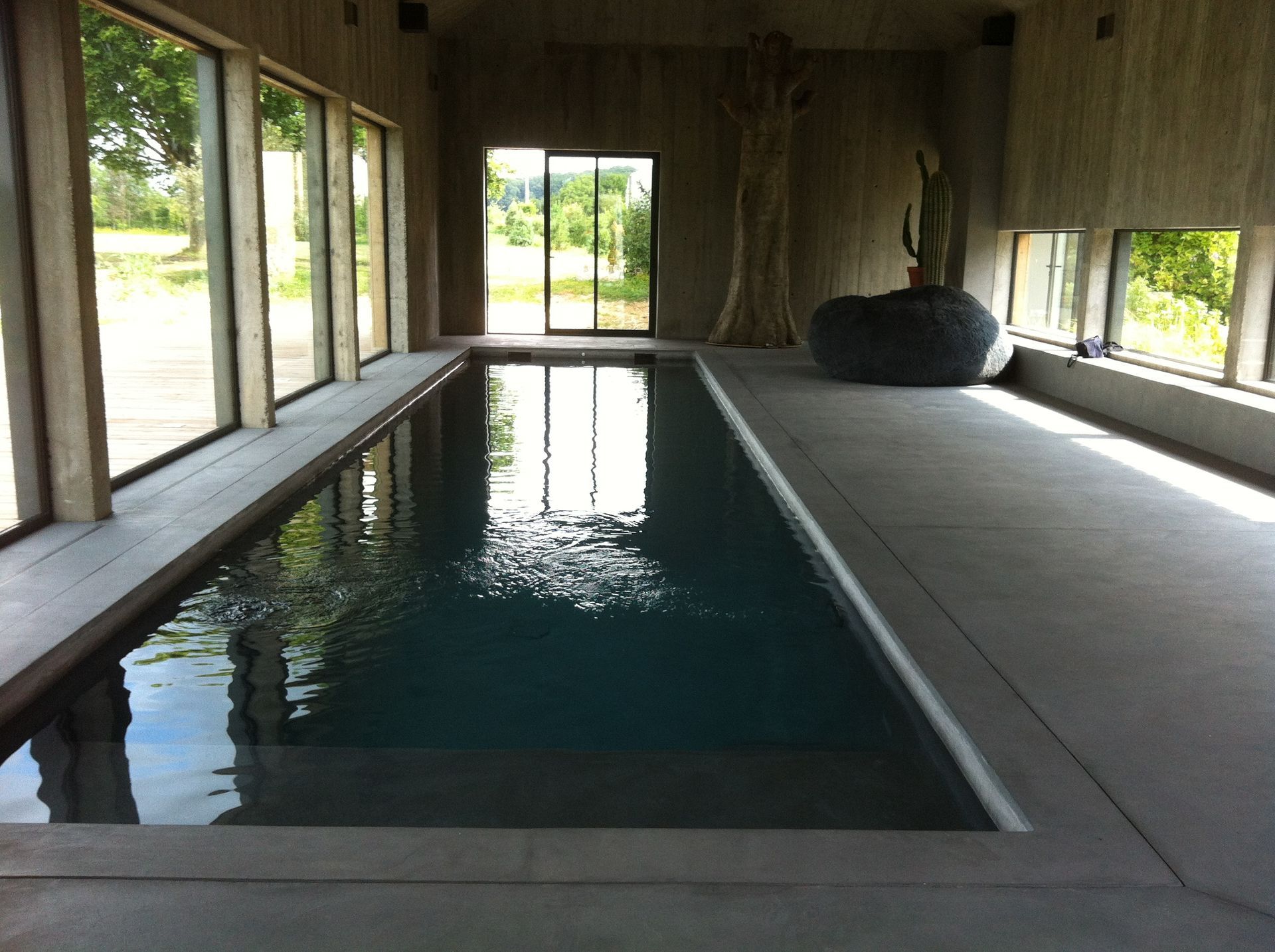 Piscine intérieure en béton avec de grandes baies vitrées donnant sur un paysage verdoyant. 