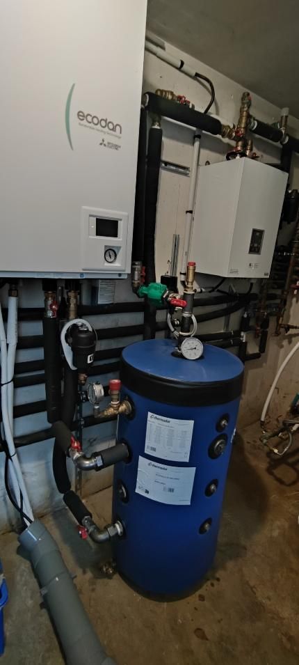 Équipements de plomberie : unité de pompe à chaleur, réservoir d’eau et tuyaux contre un mur dans un sous-sol.