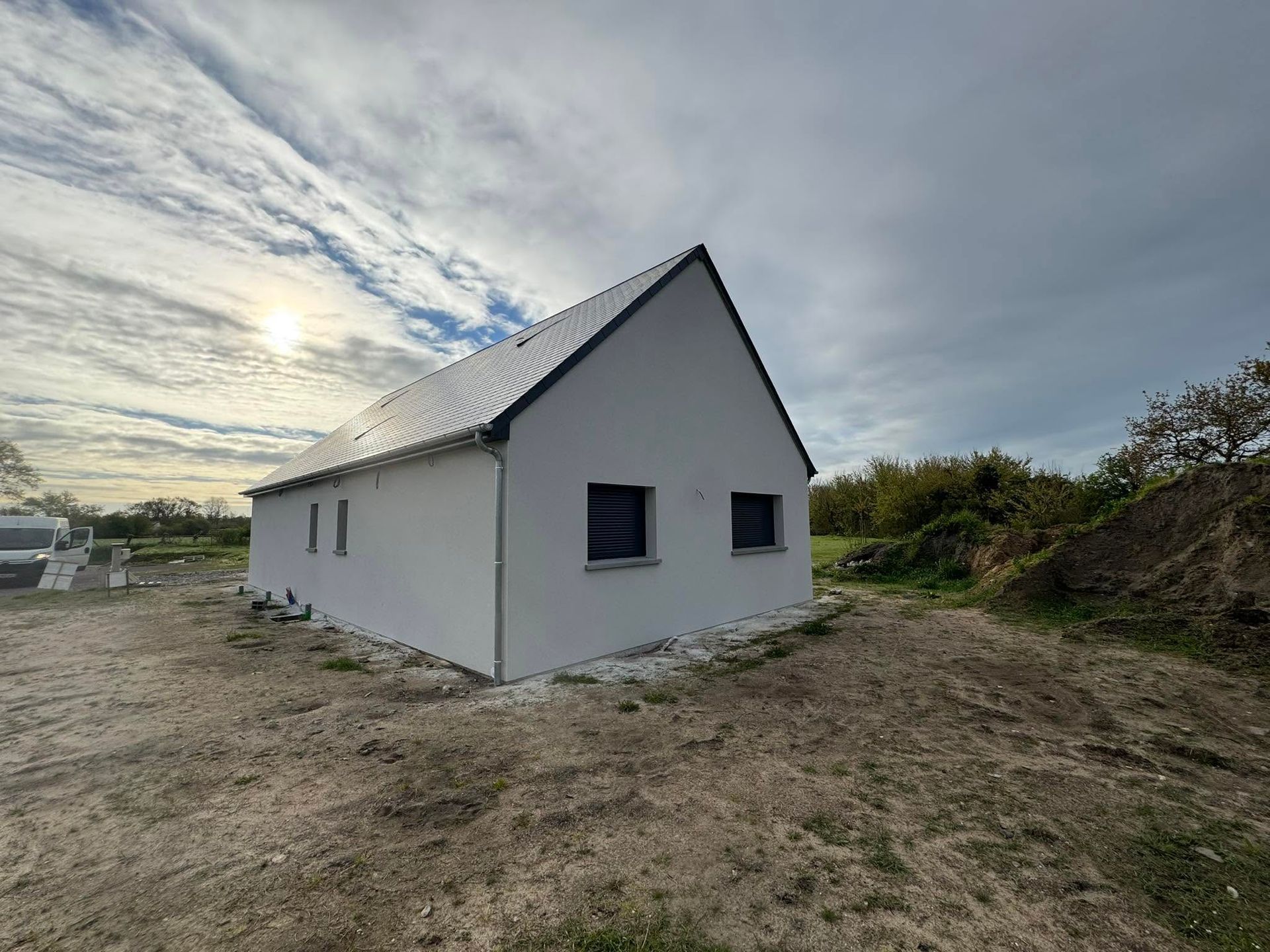Petite maison blanche au toit gris dans un champ rural sous un ciel nuageux
