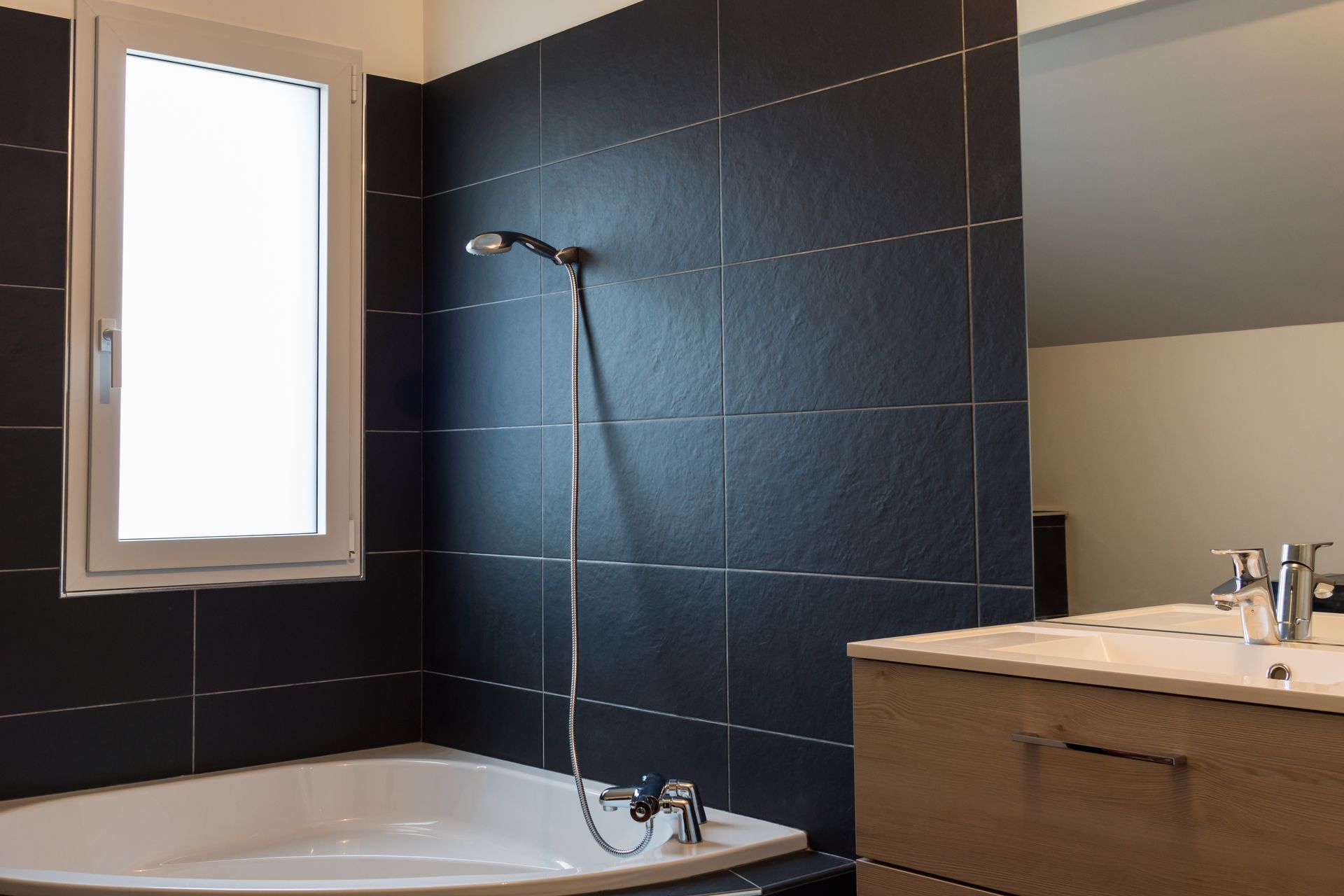 Salle de bains moderne avec carrelage noir, baignoire, pommeau de douche, fenêtre et meuble-lavabo.