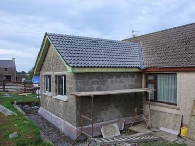 Petite extension de maison inachevée avec des murs en parpaings gris, un toit de tuiles et une fenêtre à côté d'une maison plus grande